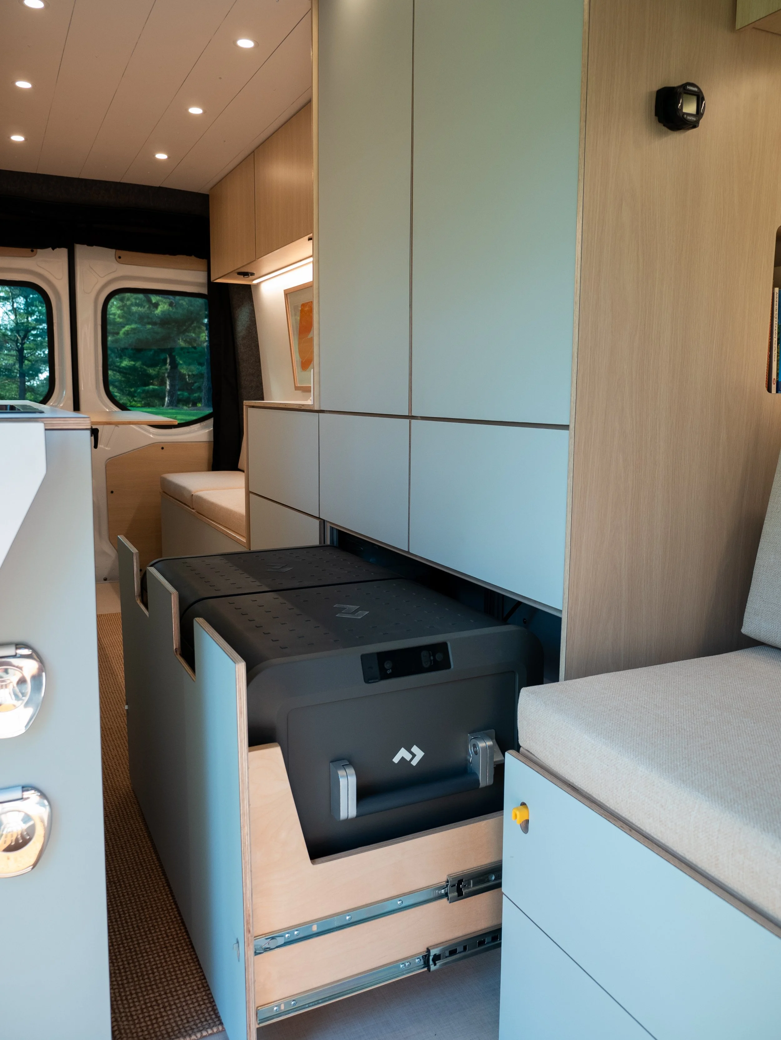 Galley and cabinetry in 'Aina' — pull-out compressor fridge on drawer slide, sage cabinetry with recessed ceiling lighting, and orange abstract artwork visible in the living area beyond