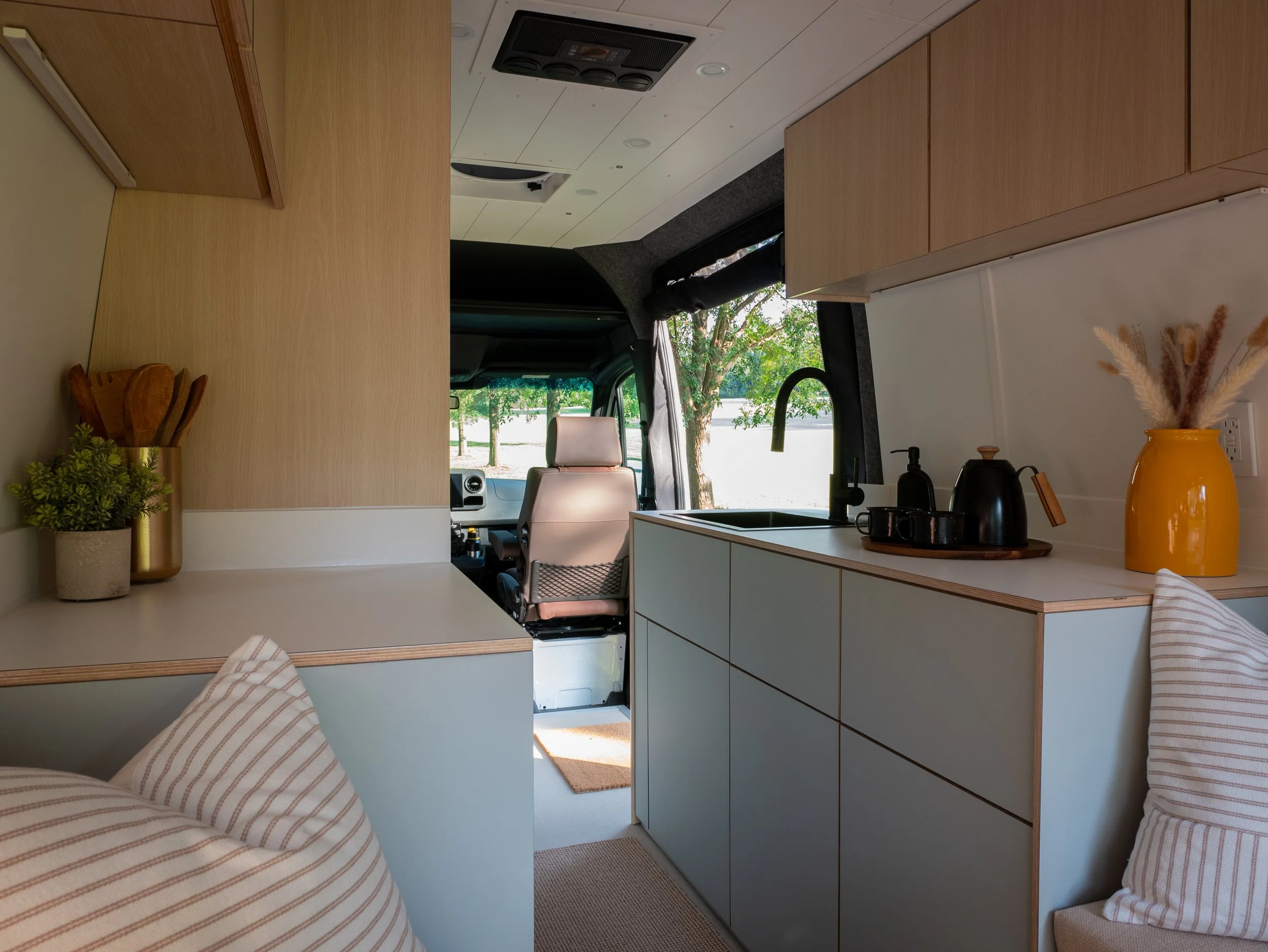 Full interior overview of 'Aina' Sprinter van conversion looking toward the cab — left counter with brass utensil holder and small plant, right galley with sage-green handleless drawers, matte black undermount sink and gooseneck faucet, black kettle 