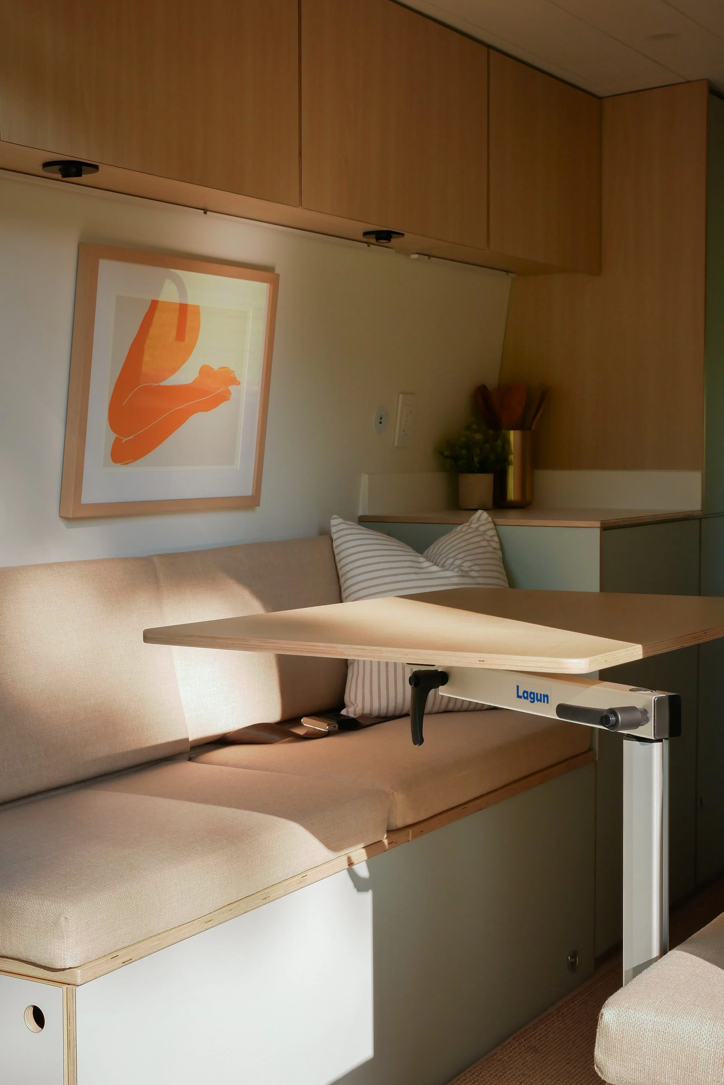 Rear lounge in 'Aina' Mercedes Sprinter van conversion — light oar bench seat cushion with striped pillow and Lagun folding table on a chrome arm; framed orange abstract artwork on the wall behind, birch plywood overhead cabinets with puck lighting, 