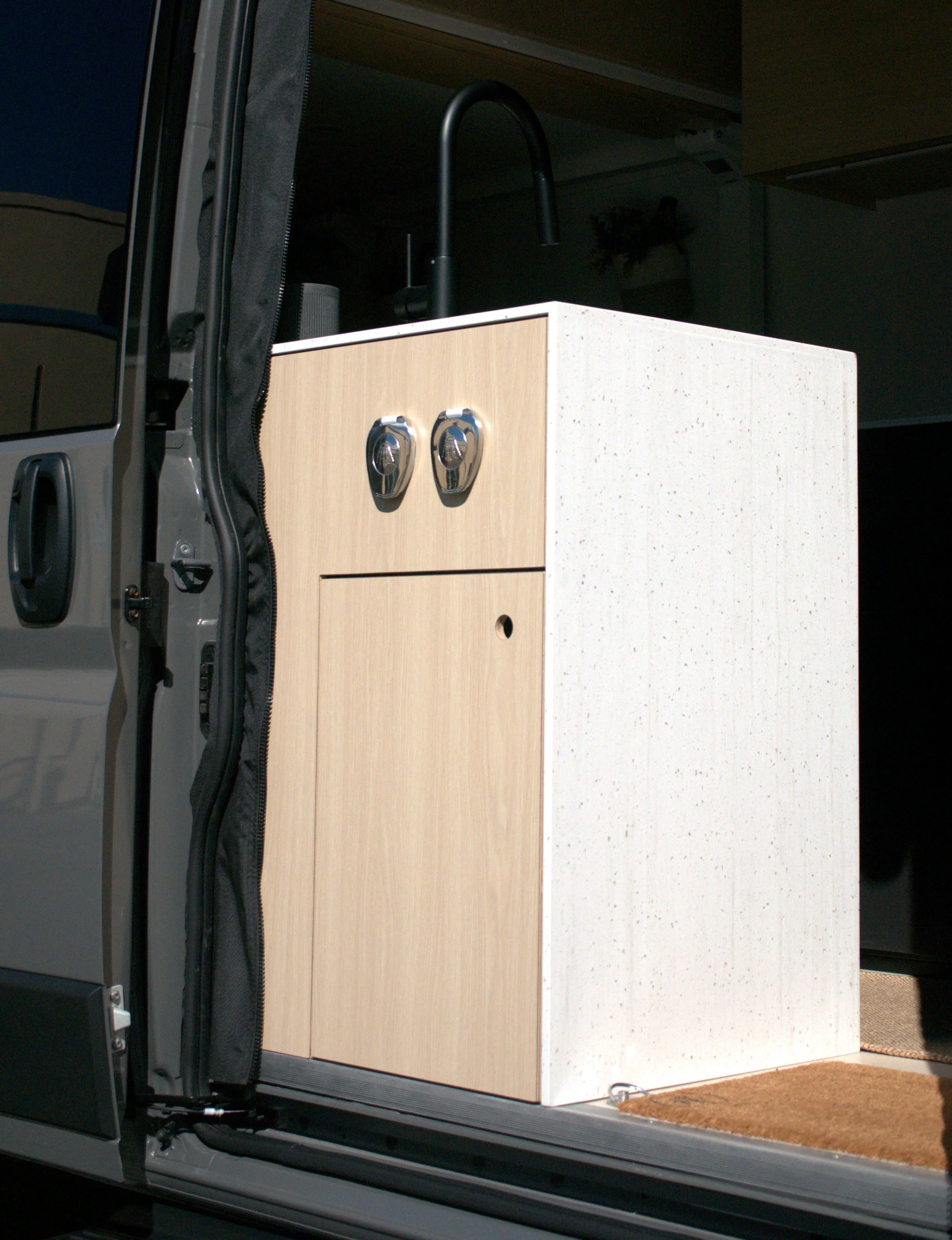 Entry kitchen module in 'Bjorn' Ram ProMaster van conversion viewed from outside through the open sliding door — light birch cabinet face with dual chrome shower port covers and routed pull, white waterfall countertop side panel, black gooseneck fauc