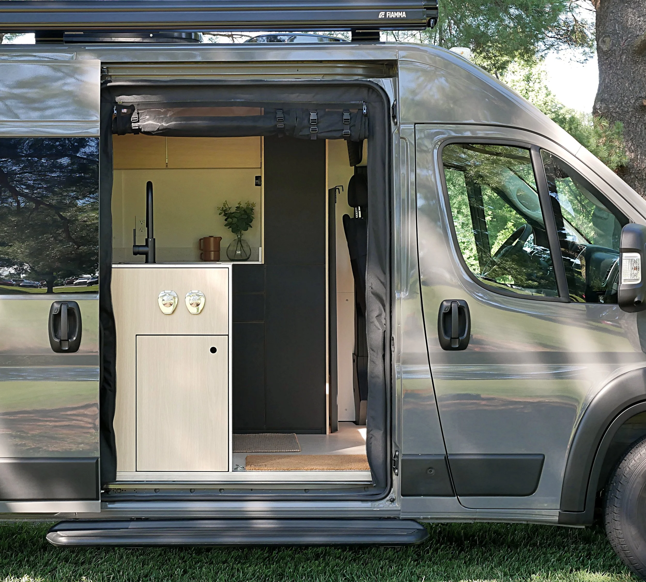'Bjorn' Ram ProMaster van conversion with sliding door open in a shaded outdoor setting — Fiamma awning overhead, birch kitchen cabinetry with outdoor shower ports at the entry, black gooseneck faucet, terracotta pot with greenery on the counter, bla
