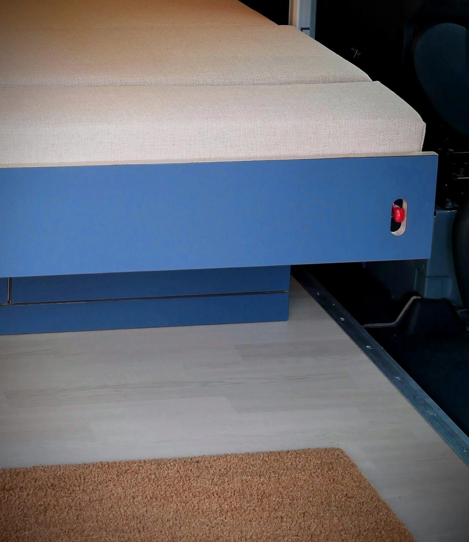 Sleeping platform bench slide-out in 'Nordlys' — blue bed panel with red release button partially extended on drawer slides, cream cushions on top, coir step mat below