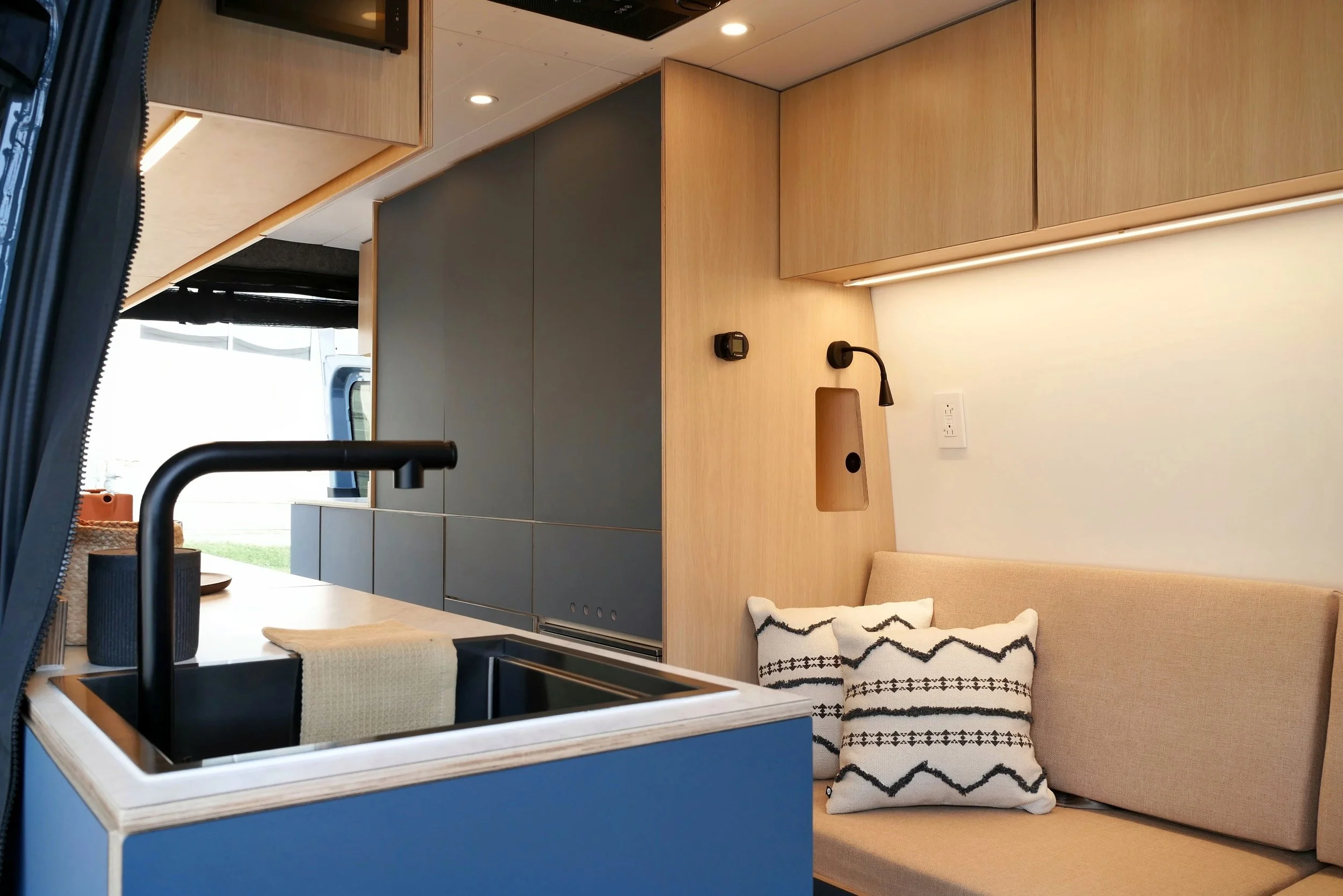 Kitchen and lounge in 'Nordlys' — blue cabinetry with black undermount sink and gooseneck faucet, bench seat with geometric black-and-white cushion, plywood ceiling with LED lighting