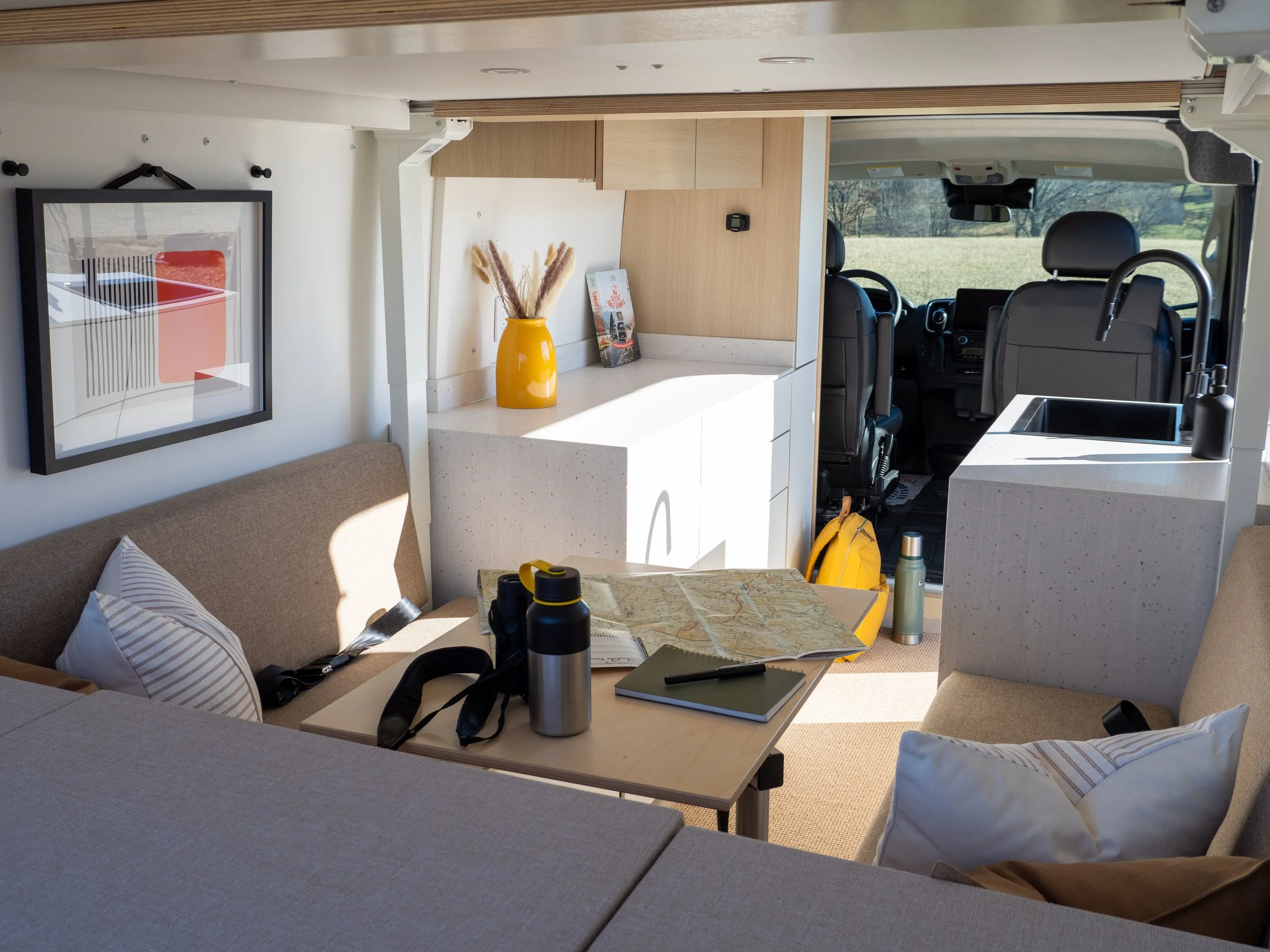 Interior lifestyle shot of 'Frida' Ram ProMaster van conversion looking from the rear toward the cab — bench seats on both sides with striped pillows, Lagun table with map, binoculars and thermos, amber vase and travel book on the kitchen counter, re