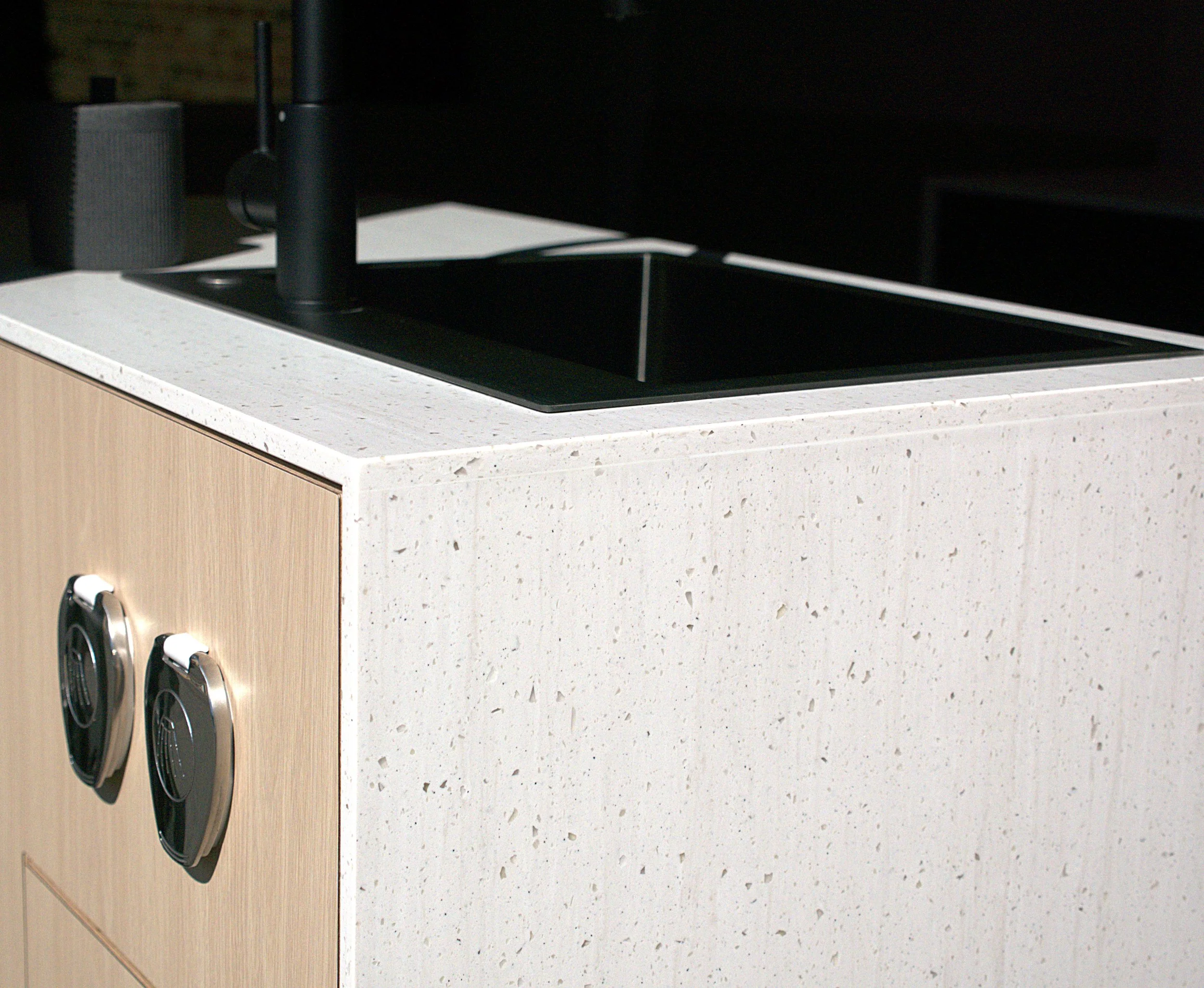 Custom van conversion kitchen in 'Bjorn' — white speckled waterfall countertop with black sink, black gooseneck faucet, r, and light birch cabinet face with polished chrome shower port covers.