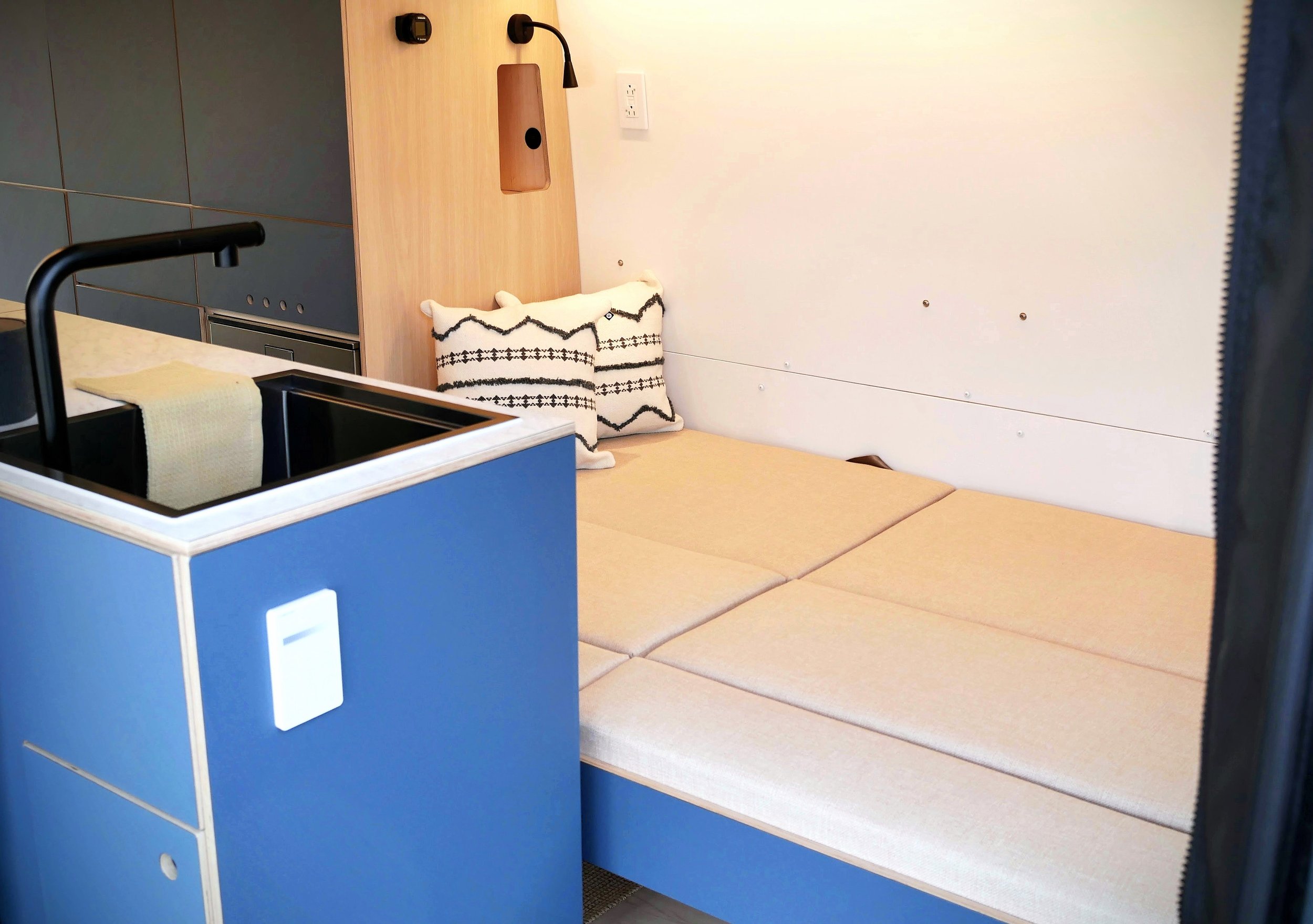 Kitchen and sleeping area in 'Nordlys' — blue galley sink unit with black sink and light dimmer unit in foreground, full sleeping platform with geometric cushion and reading lamp visible behind