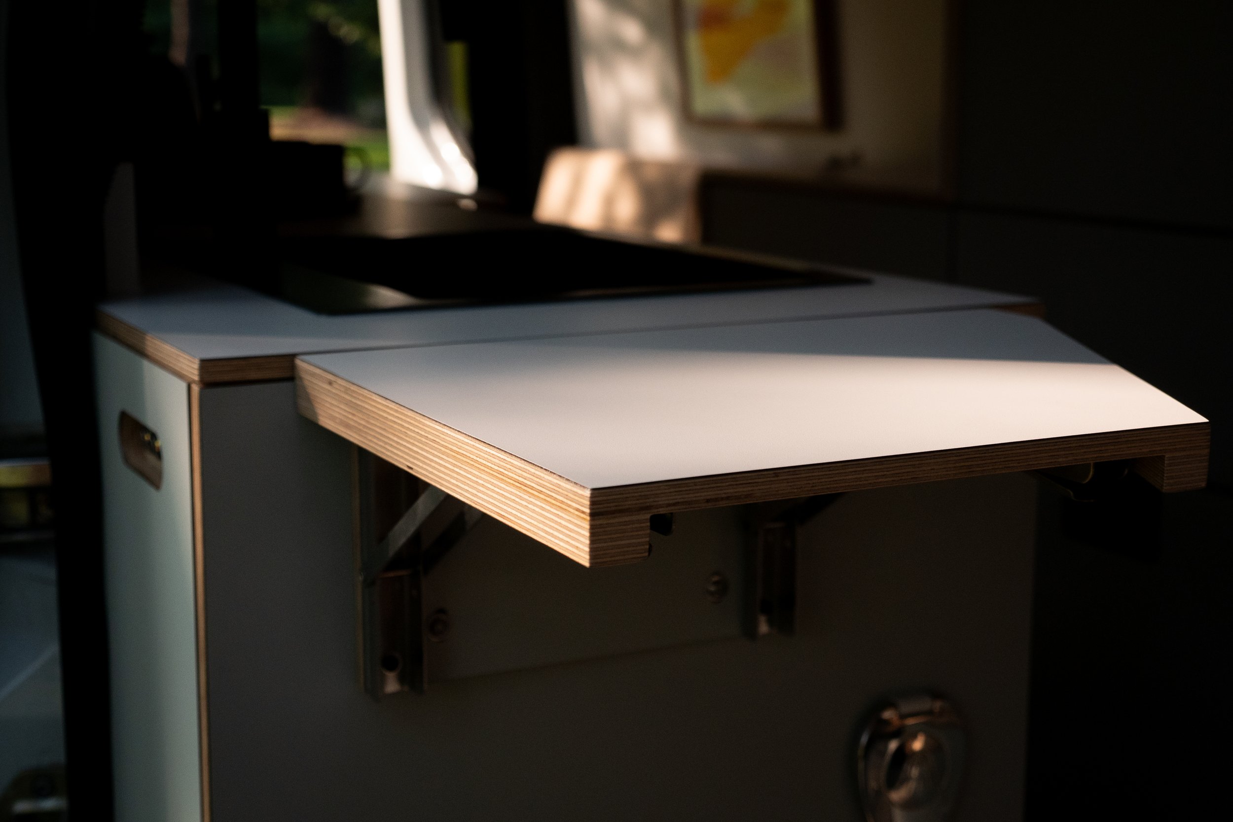 Fold-out counter extension in 'Aina' van conversion — white laminate workspace with exposed birch plywood edge, matte cabinet body with chrome latch, black metal sink above, warm raking light across the surface.