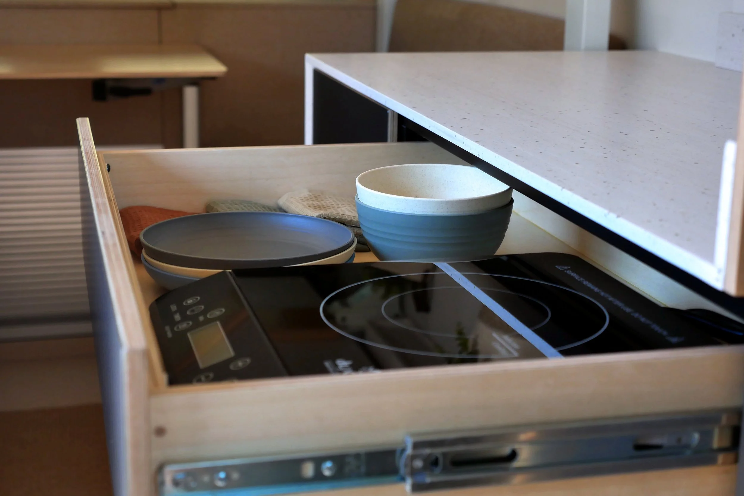 Hidden pull-out induction cooktop in 'Bjorn' Ram ProMaster van conversion — large birch plywood drawer on full-extension slides reveals a two-burner induction hob with digital controller, stacked charcoal plates, blue ceramic bowl, and linen cloth; w
