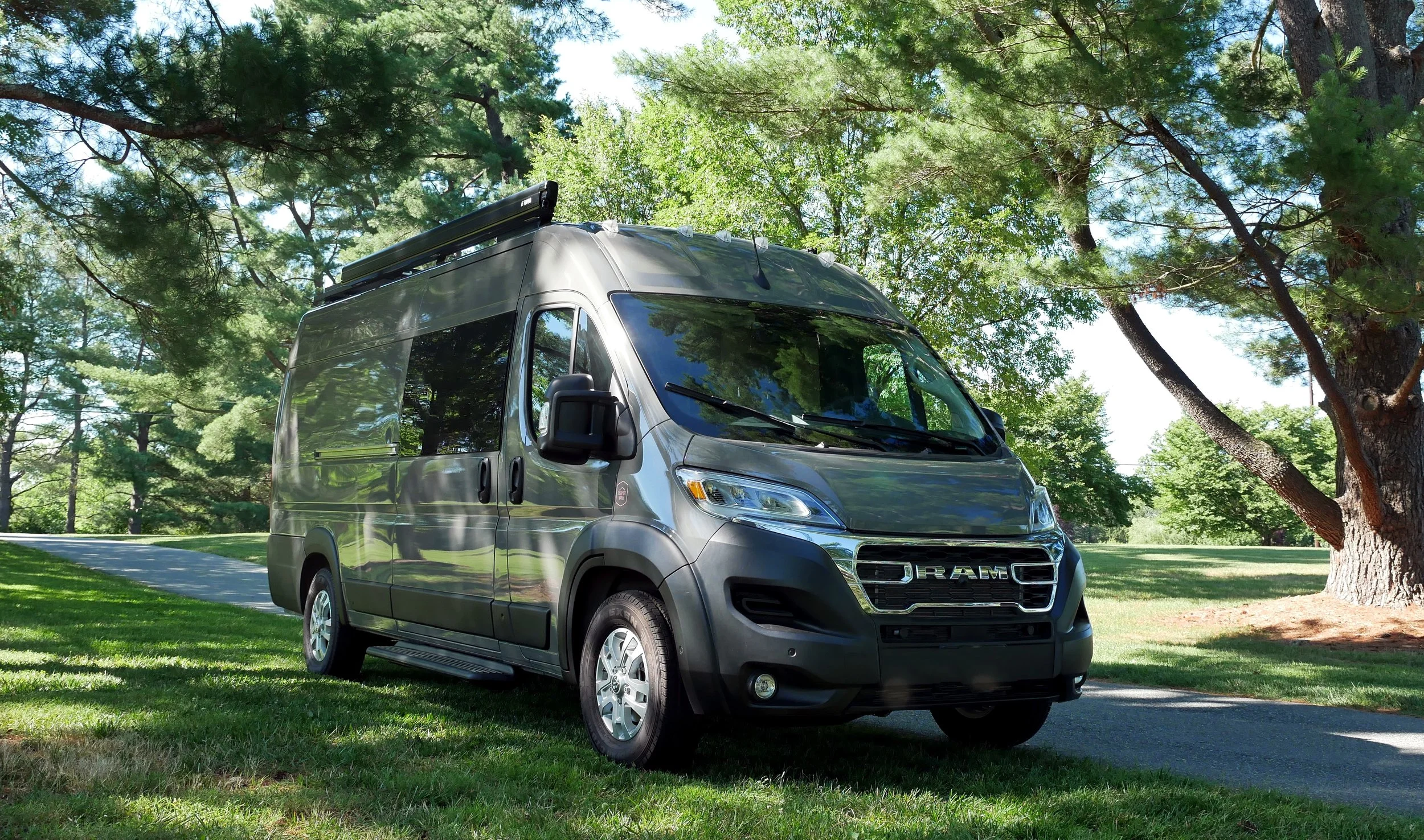 Full exterior view of 'Bjorn' Ram ProMaster high-roof van conversion — grey ProMaster parked on a grassy lawn under tall pine trees, black roof rack with solar panels, chrome RAM front badge, tinted side windows in dappled afternoon light.