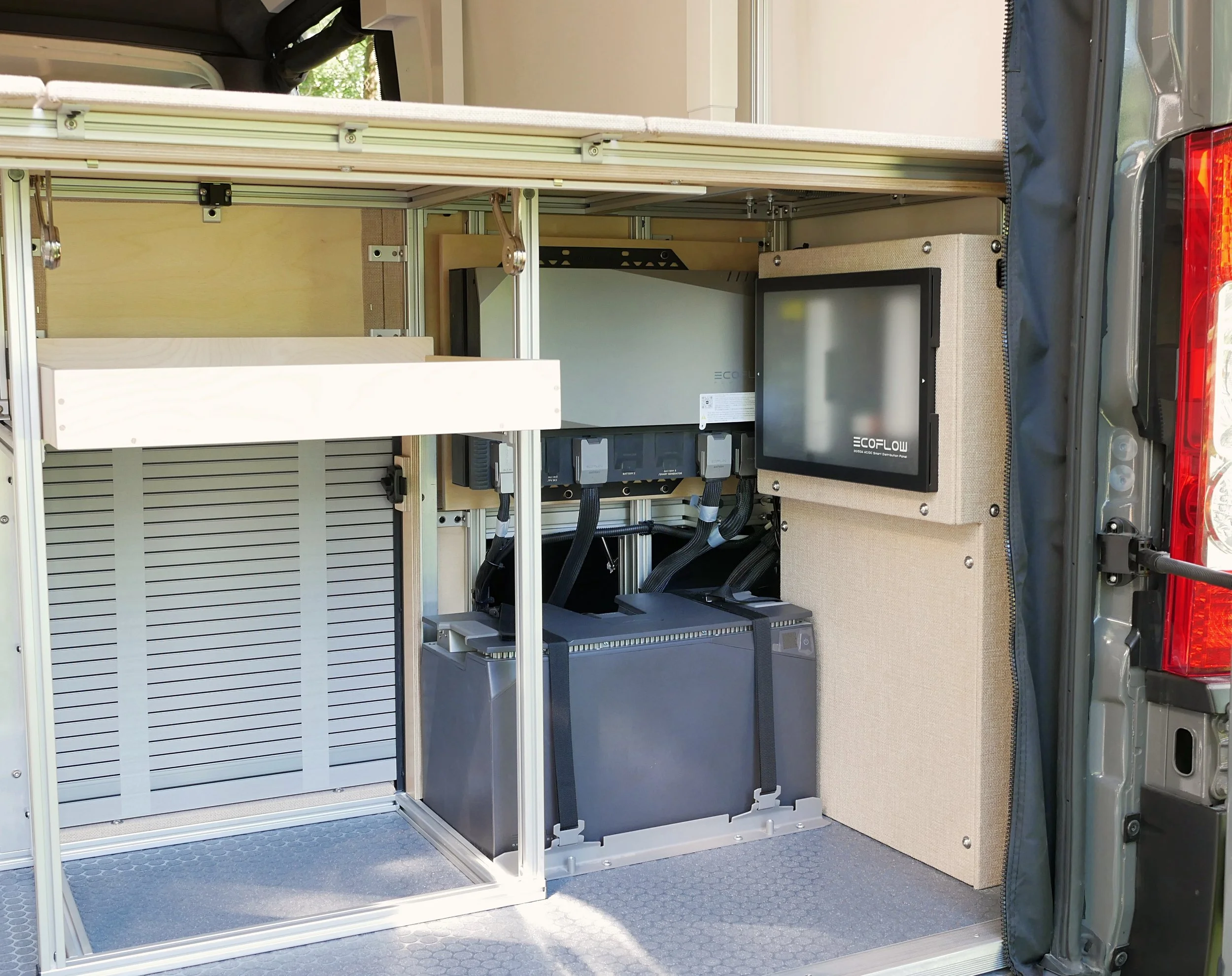 Rear electrical and gear bay of 'Bjorn' Ram ProMaster van conversion fully open — louvered storage panel alongside aluminum-framed twin EcoFlow lithium battery unit, EcoFlow distribution panel on the beige wall panel, Loncoin vinyl floor, and ProMast