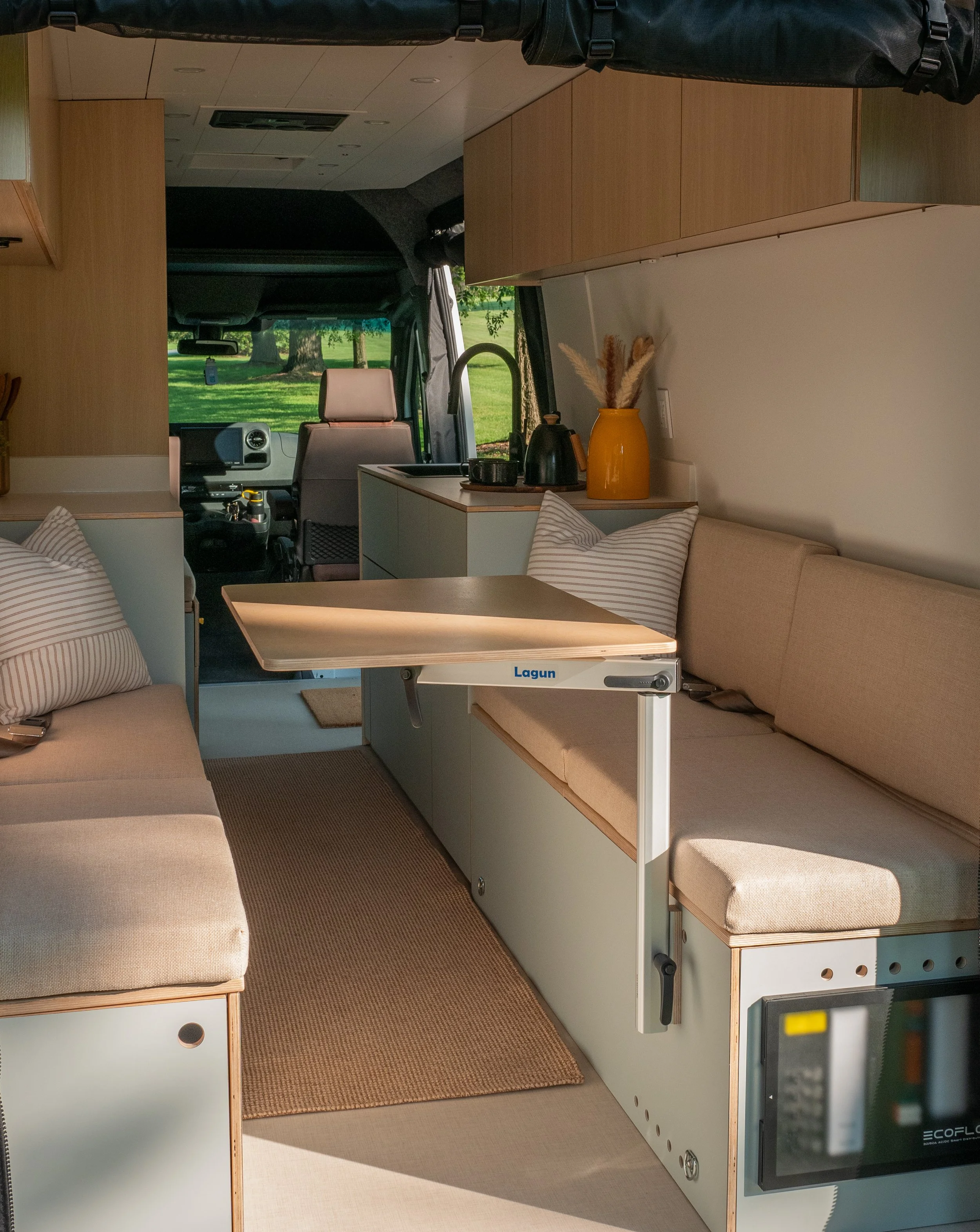 Full interior of 'Aina' Sprinter van conversion looking toward the cab — facing light oat upholstered bench seats on sage-green cabinet bases with striped pillows and a Lagun folding table, woven jute runner, EcoFlow distribution panel at the bench b