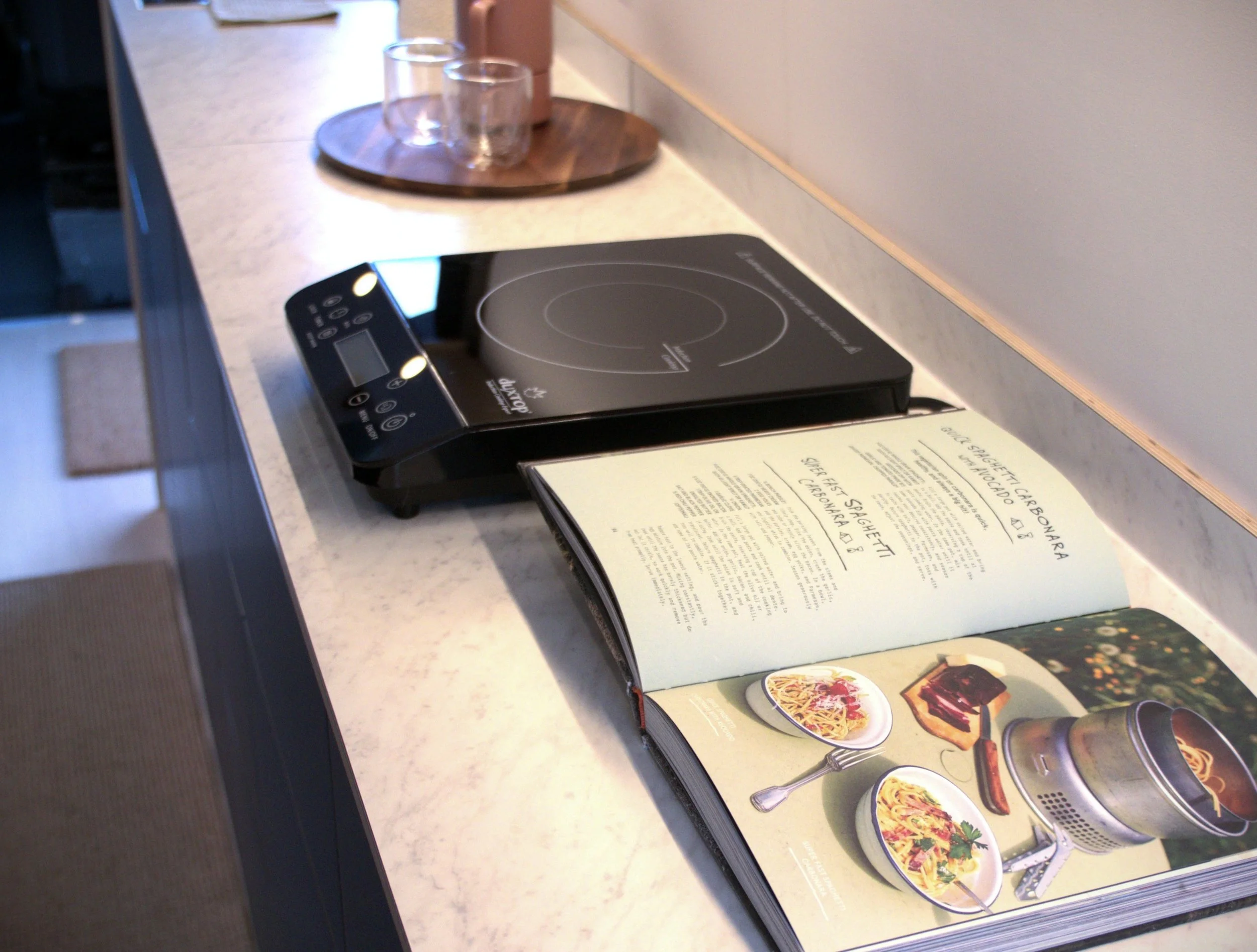 Induction cooktop lifestyle detail in 'Nordlys' — portable burner on white countertop, open cookbook beneath it, copper tray and glassware, LED under-cabinet lighting