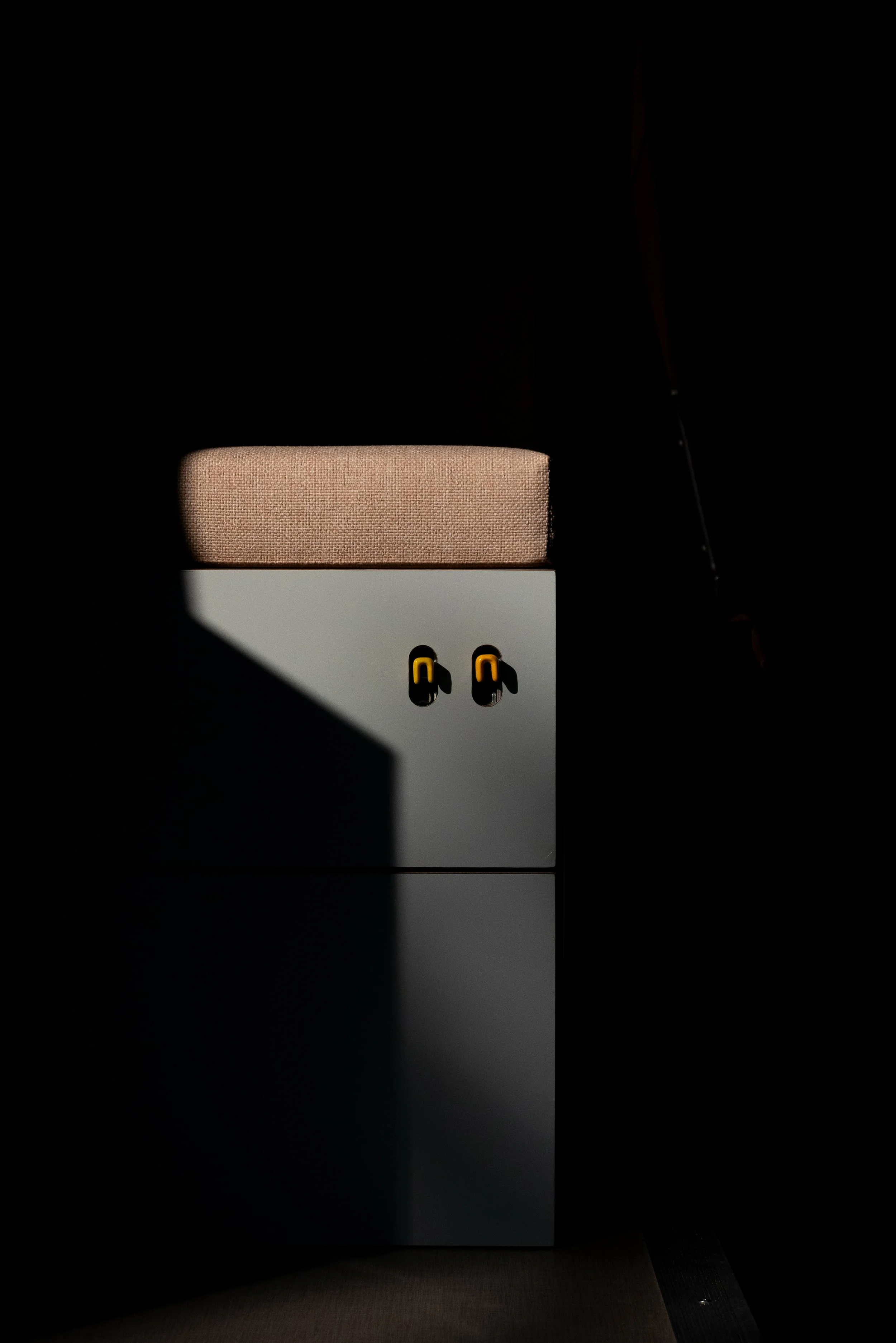 Custom van conversion hardware detail in 'Aina' — convertible bench seat with twin heavy duty drawer slide tabs illuminated by a single shaft of sunlight, topped with a natural jute-upholstered cushion pad, deep shadow framing the composition.