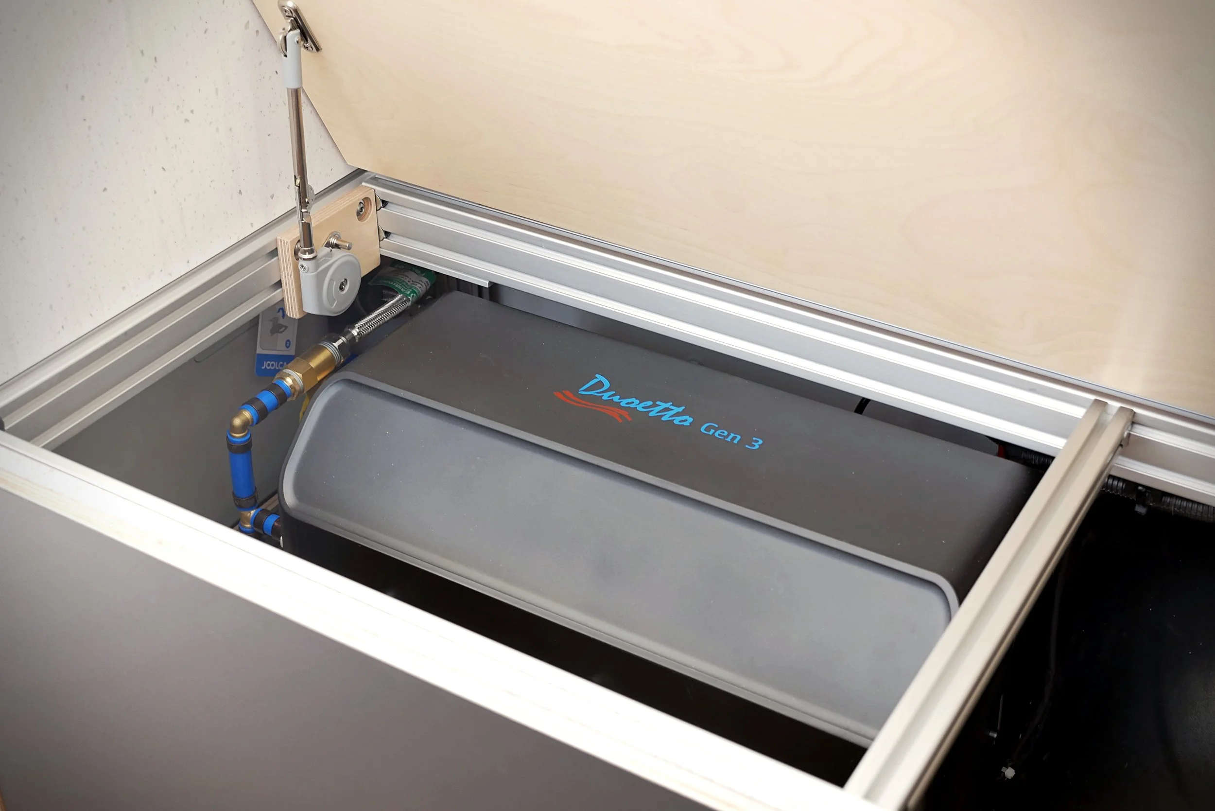 Van conversion hot water system in 'Bjorn' — aluminum-framed bench seat hatch open revealing the Duoetto Gen 3 water heater with blue and red branding, plumbing connections at left, strut prop holding the hatch open.