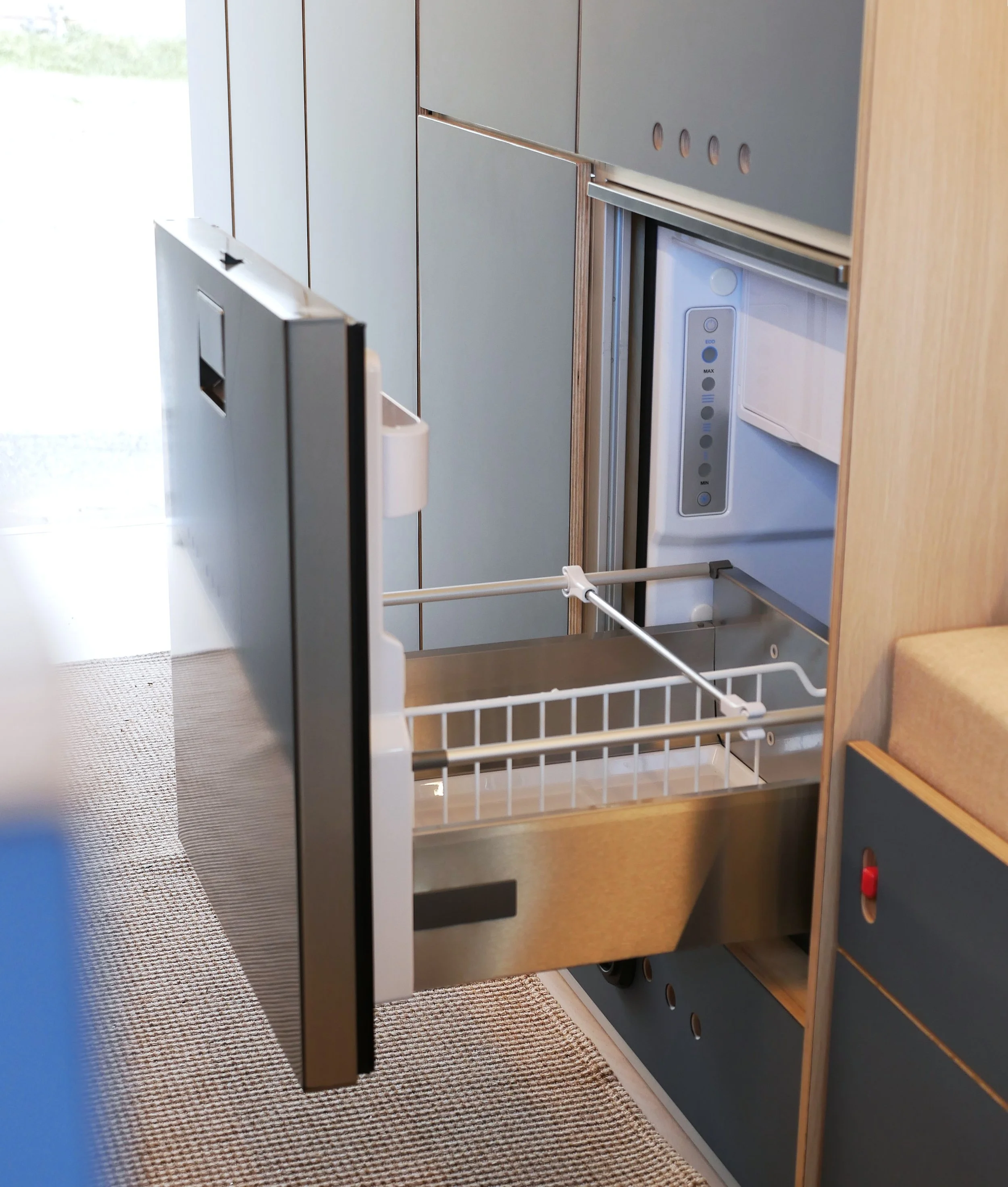 Off Indel compressor fridge open in 'Nordlys' — stainless interior with wire basket, tall black door swung open, blue cabinetry surrounding, woven rug floor