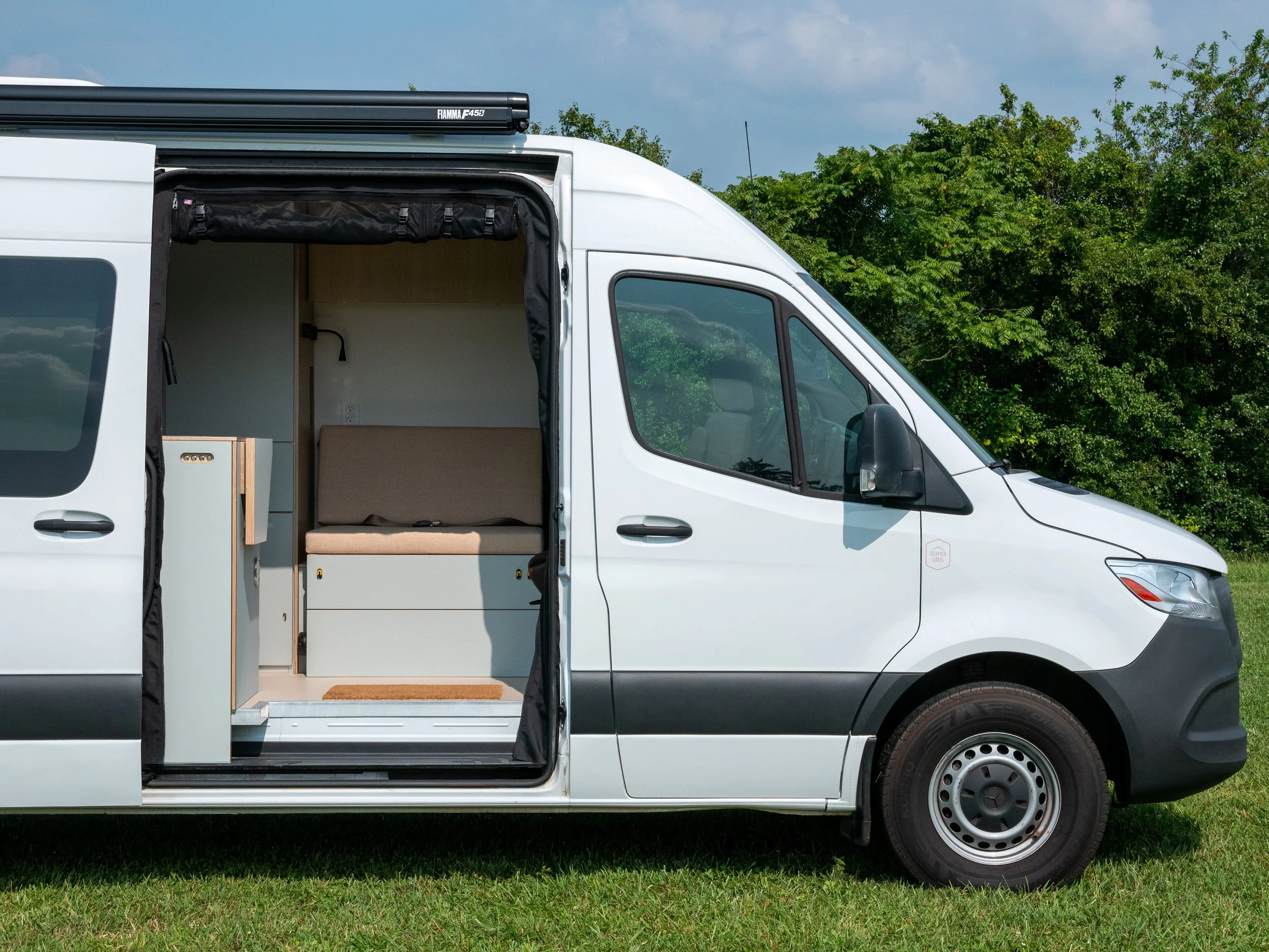 White Mercedes-Benz Sprinter van conversion 'Aina' parked on a grassy lawn with the sliding door open — a custom Maryland campervan build with sage-green lower cabinetry, tan bench cushion and coir entry mat visible through the open door, Fiamma F450