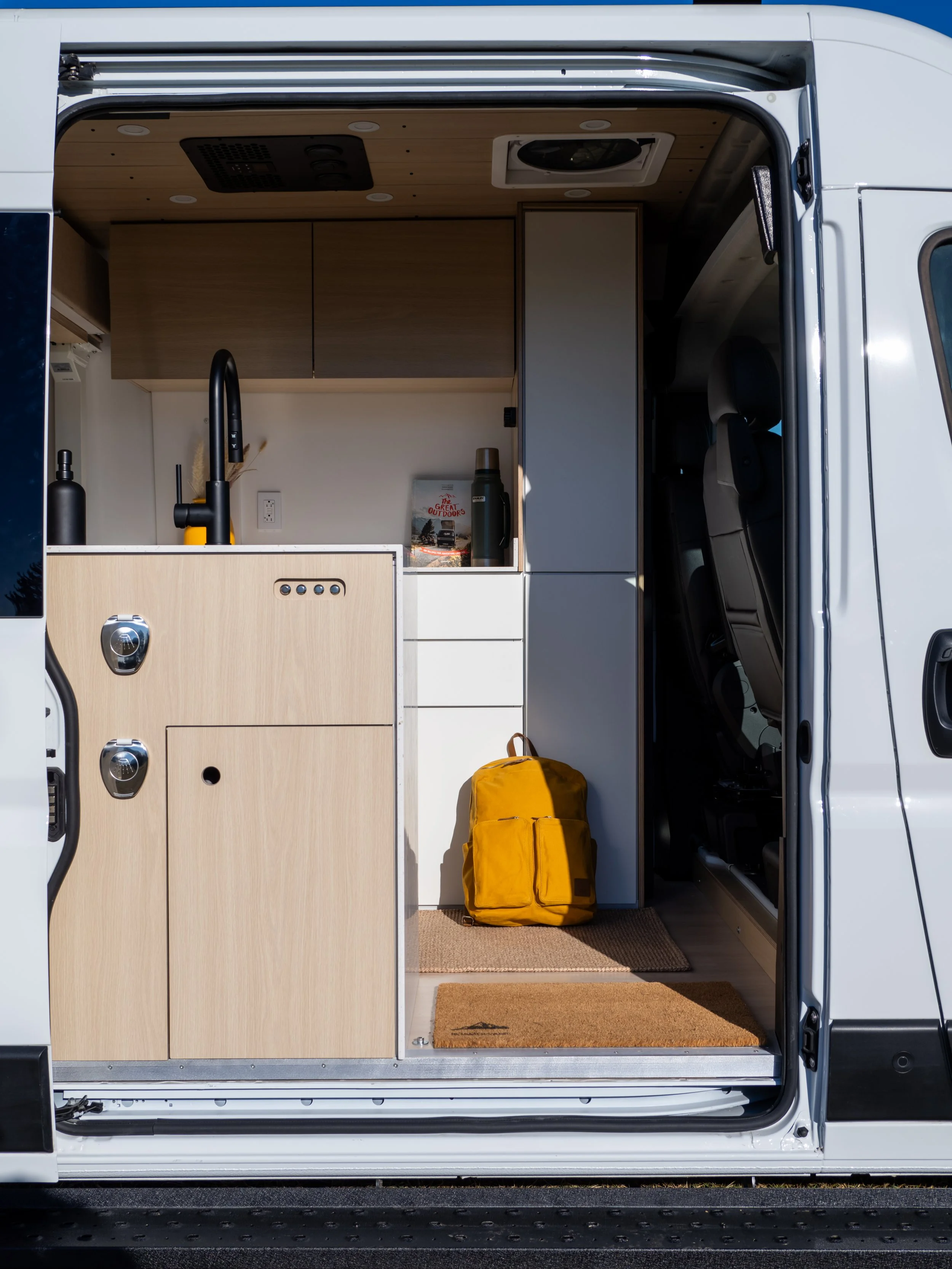 View through the open sliding door into 'Frida' Ram ProMaster van conversion — birch plywood cabinetry and black gooseneck faucet on the left, grey overhead cabinets, MaxxFan ceiling vent, yellow backpack on the coir entry mat.