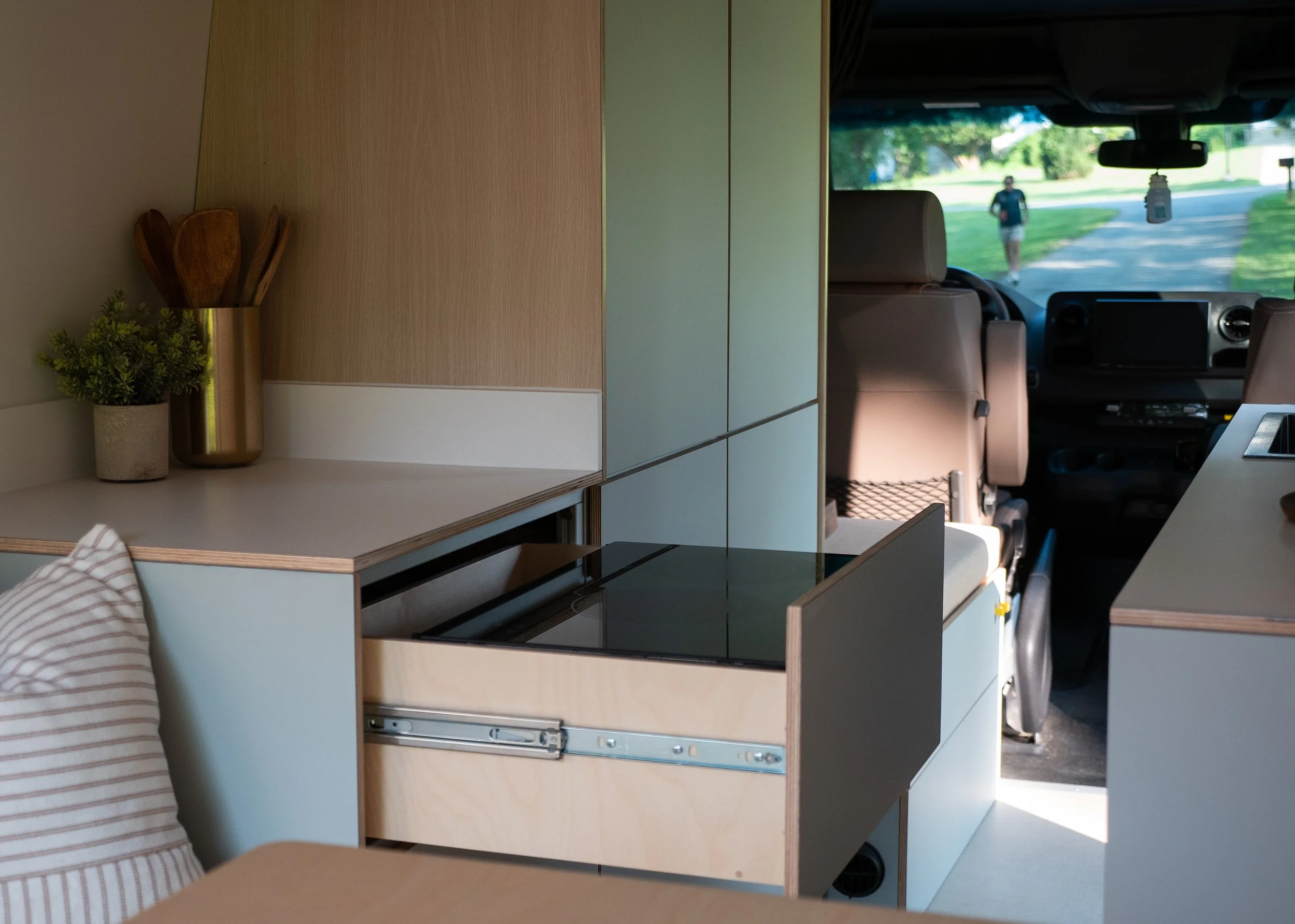 Hidden pull-out induction cooktop in 'Aina' Mercedes Sprinter van conversion — birch plywood drawer on full-extension slides reveals a flush black induction hob beneath the white countertop, sage-green flat-front cabinet face below; brass utensil hol