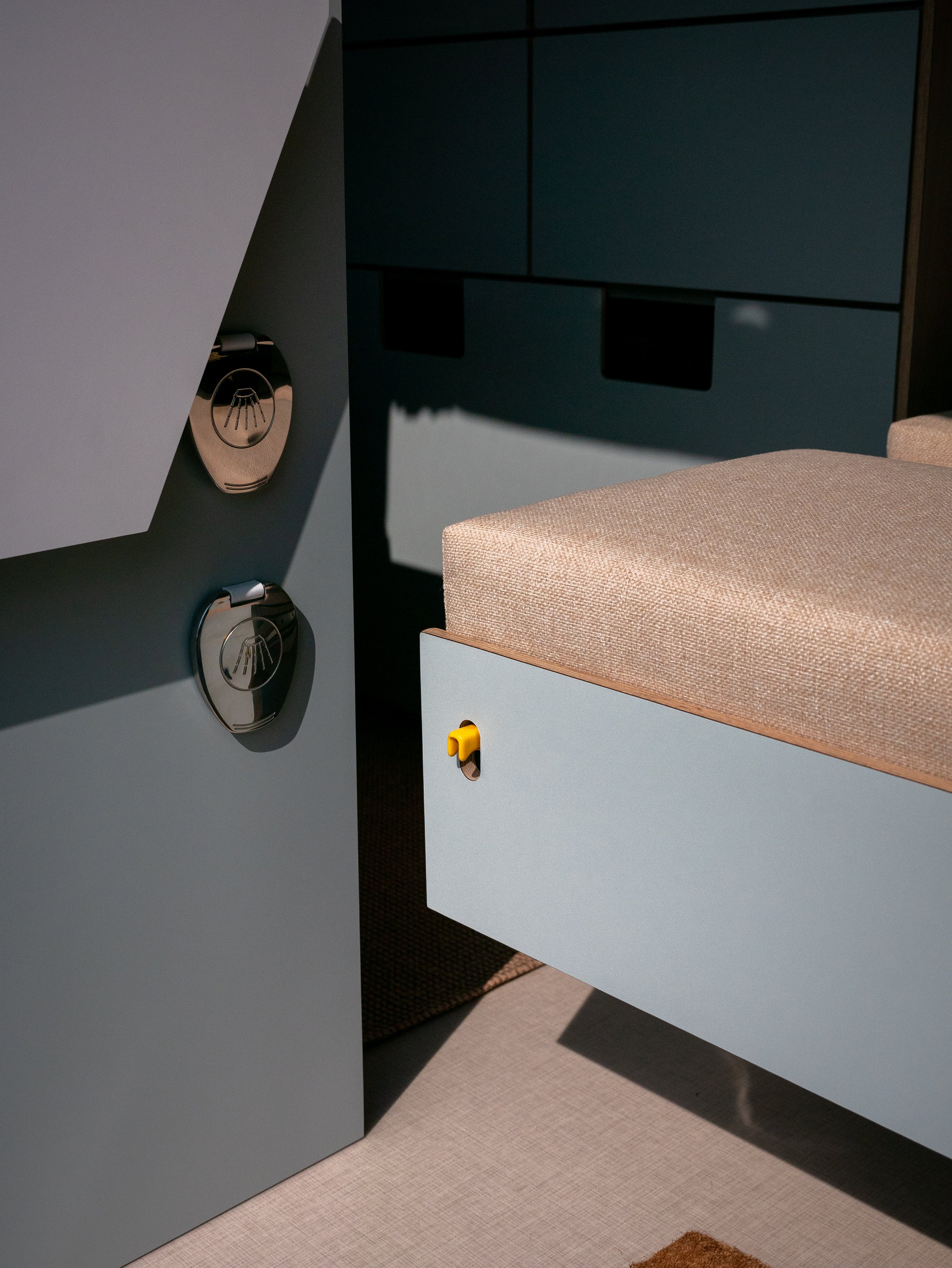 Van conversion hardware close-up in 'Aina' — twin polished chrome shower ports , sage-green cabinetry and a natural linen-upholstered step bench with yellow latch tab visible in the background.