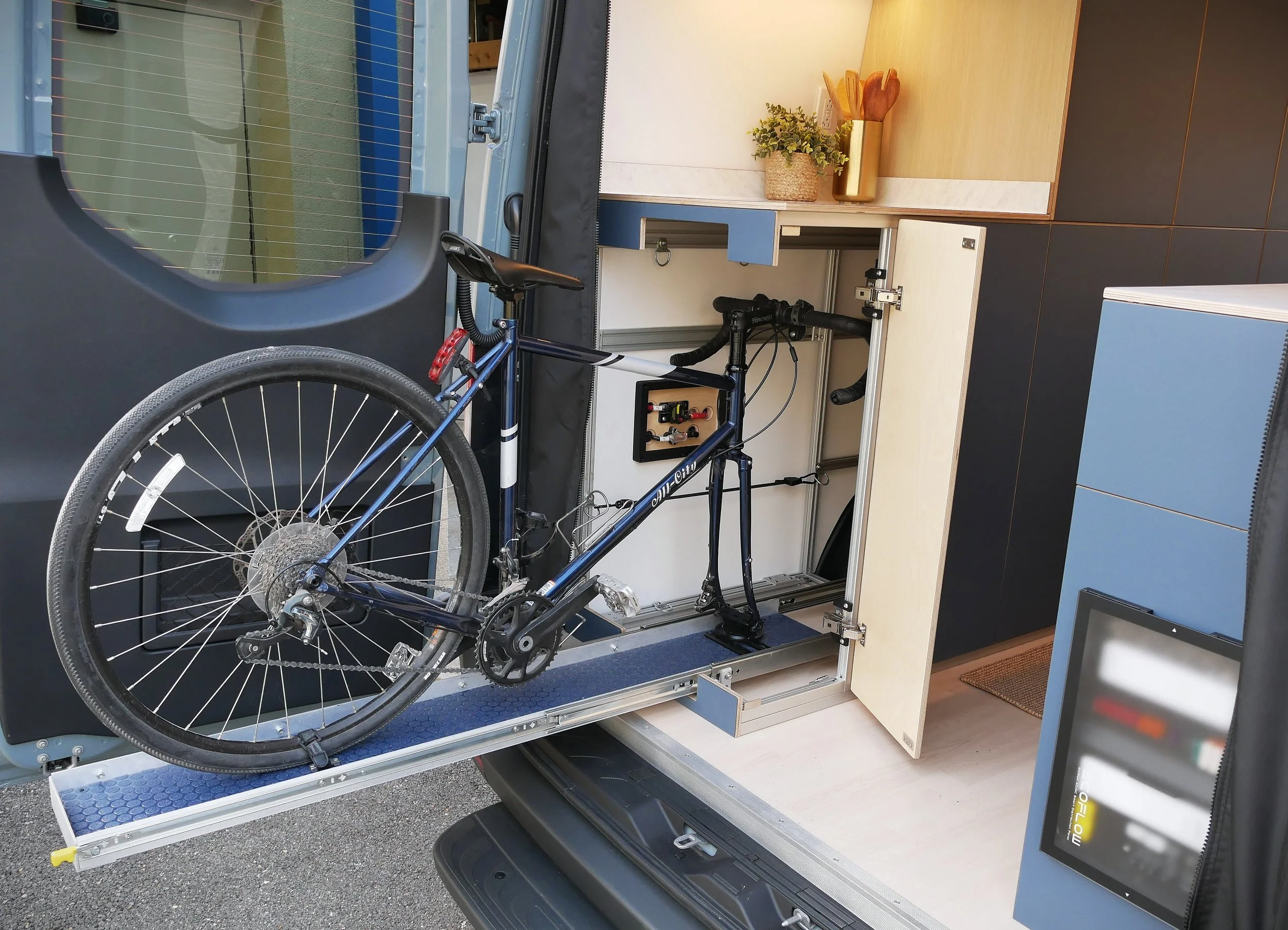 Integrated bicycle storage in 'Nordlys' — full bicycle on a custom slide-out tray extending from the rear of the van, kitchen counter with utensil holder visible inside, Ecoflow Powerkit distribution panel on right