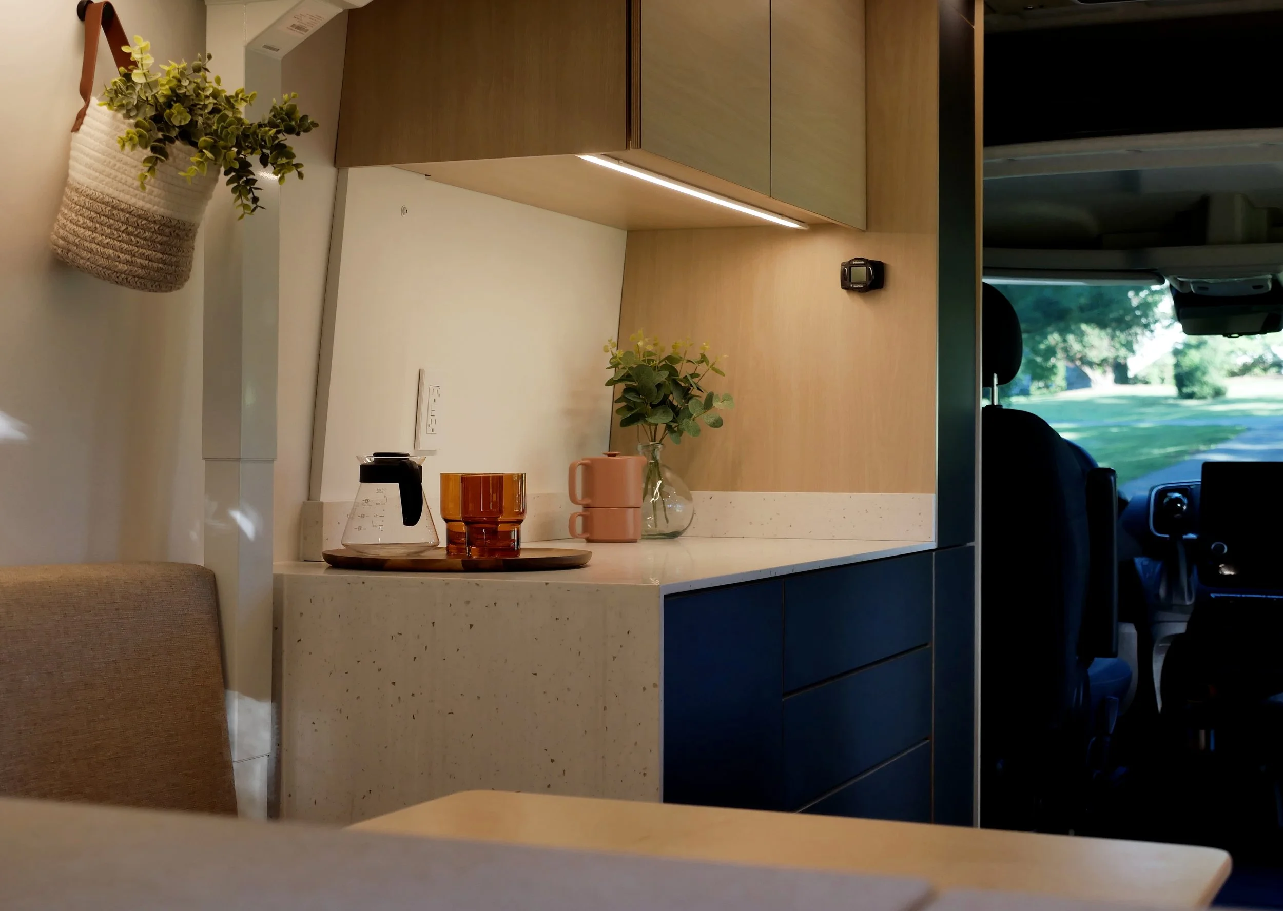 Galley kitchen in 'Bjorn' van conversion — white countertop over black flat-front handleless drawers, warm under-cabinet LED strip, birch upper cabinets; Chemex coffee maker, amber glass, pink ceramic mugs and eucalyptus styling; hanging woven plant 