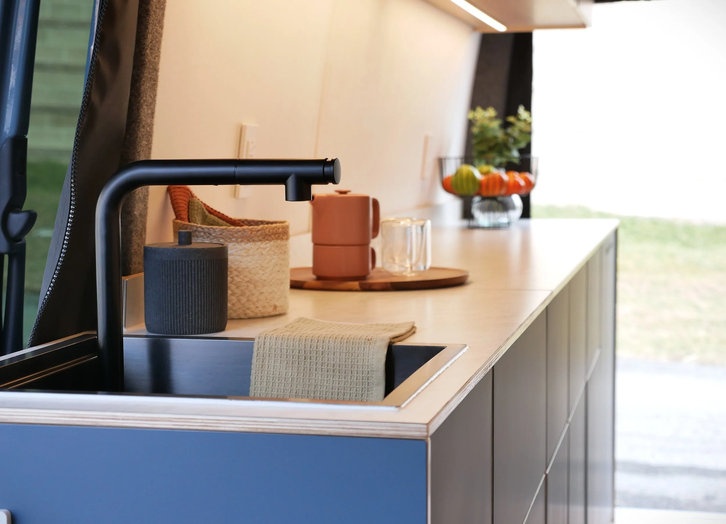 Kitchen counter in 'Nordlys' — black pull-out faucet, plywood-edged white countertop, blue cabinet face, linen dish towel, woven basket and terracotta mugs on wooden tray, LED under-cabinet lighting