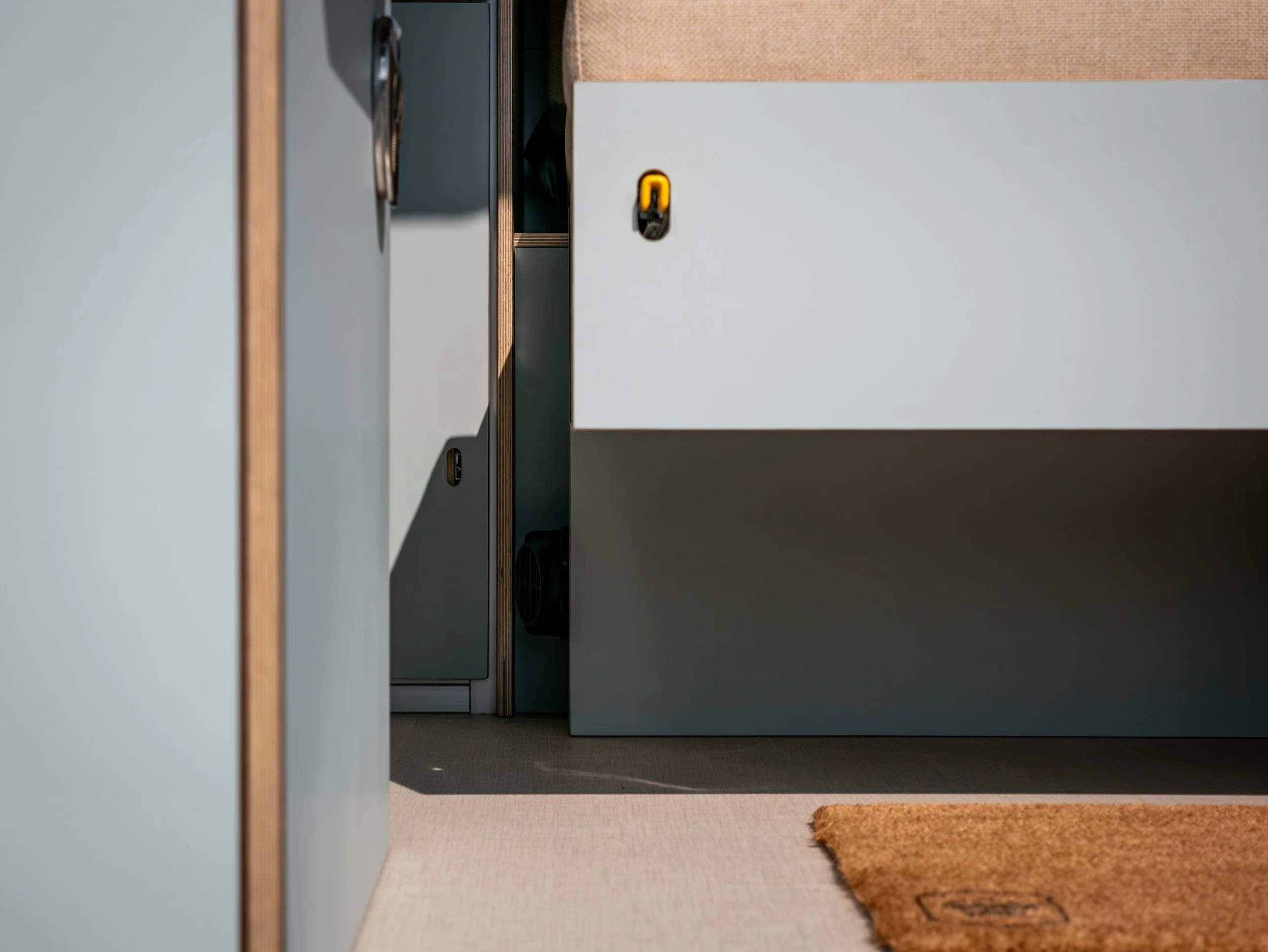 Entry threshold of 'Aina' viewed through the open sliding door — coir step mat, sage cabinetry, and pull-out platform for convertible bench seat