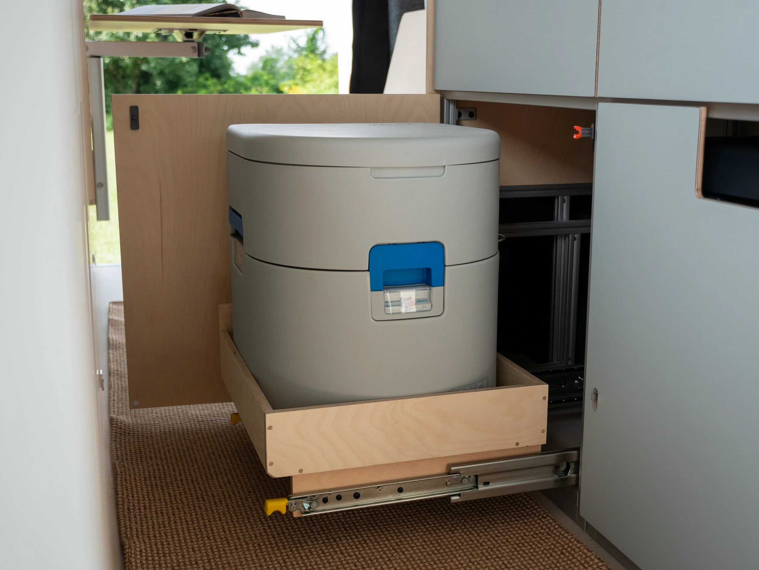 Hidden separator toilet in 'Aina' van conversion — Joolca GottaGo on a floor-level birch plywood pull-out tray on full-extension drawer slides, yellow safety latch at the base; sage-green cabinetry on either side, woven jute runner, and green trees t