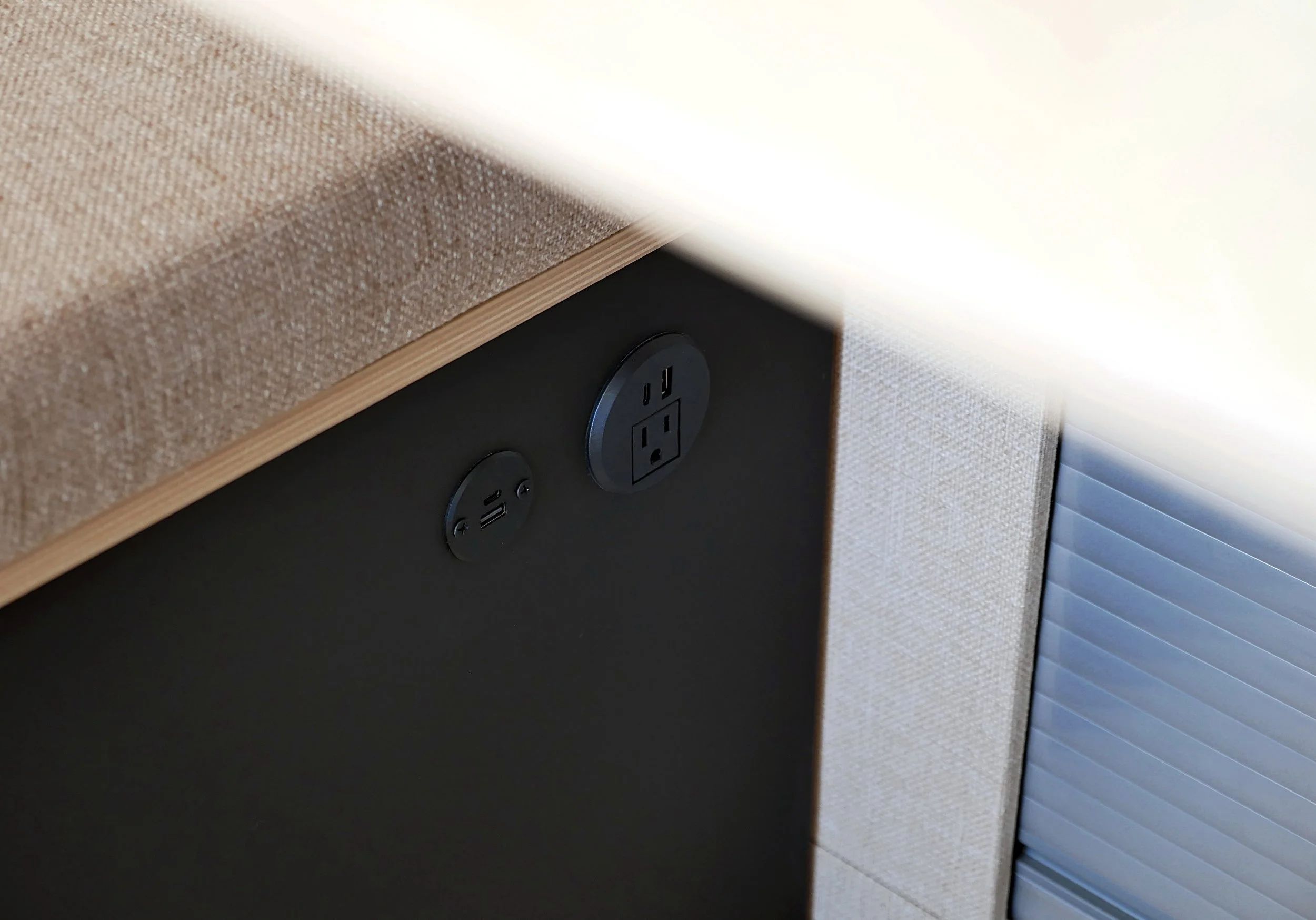 Van conversion power outlets in 'Bjorn' — flush-mounted round black USB charging port and 120V AC duplex outlet integrated into the dark bench panel below the cushion, dramatic diagonal light beam across the composition.