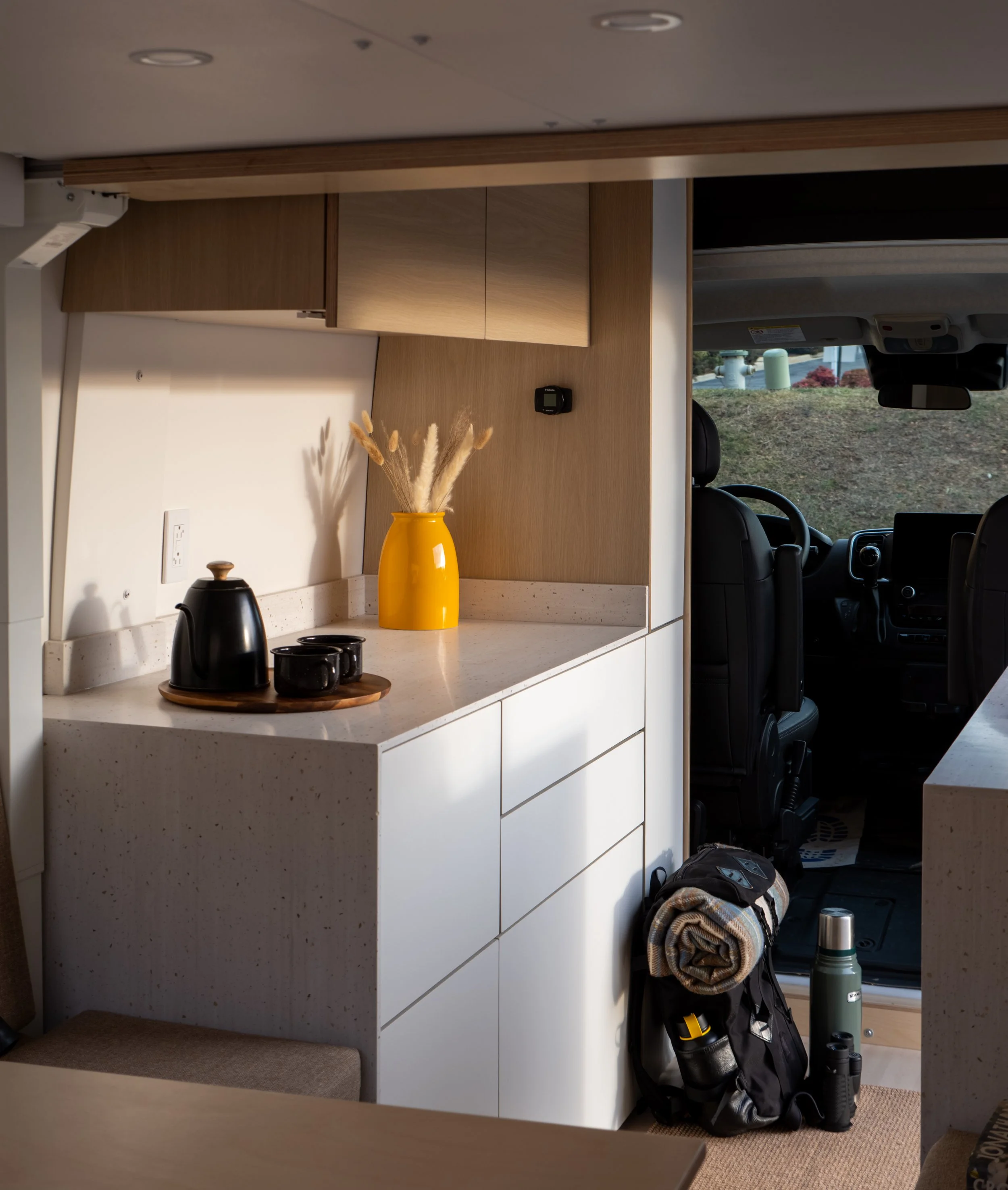 Van conversion kitchen in 'Frida' looking toward the cab — white speckled quartz countertop with black kettle on a wooden tray and two black ceramic mugs, amber pampas vase; white flat-front handleless drawer stack, birch plywood wall panel and overh