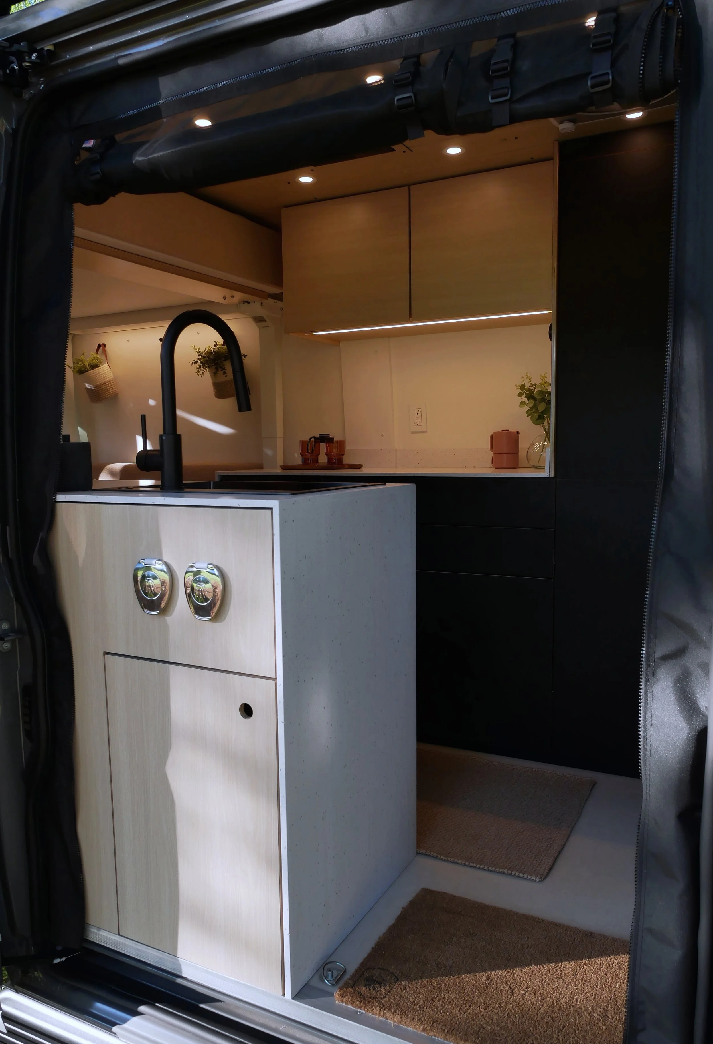 'Bjorn' Ram ProMaster van conversion interior at dusk viewed through the open sliding door — entry kitchen module with birch cabinet and chrome Southco latches in the foreground, lit galley kitchen with LED under-cabinet strip, hanging plant basket, 