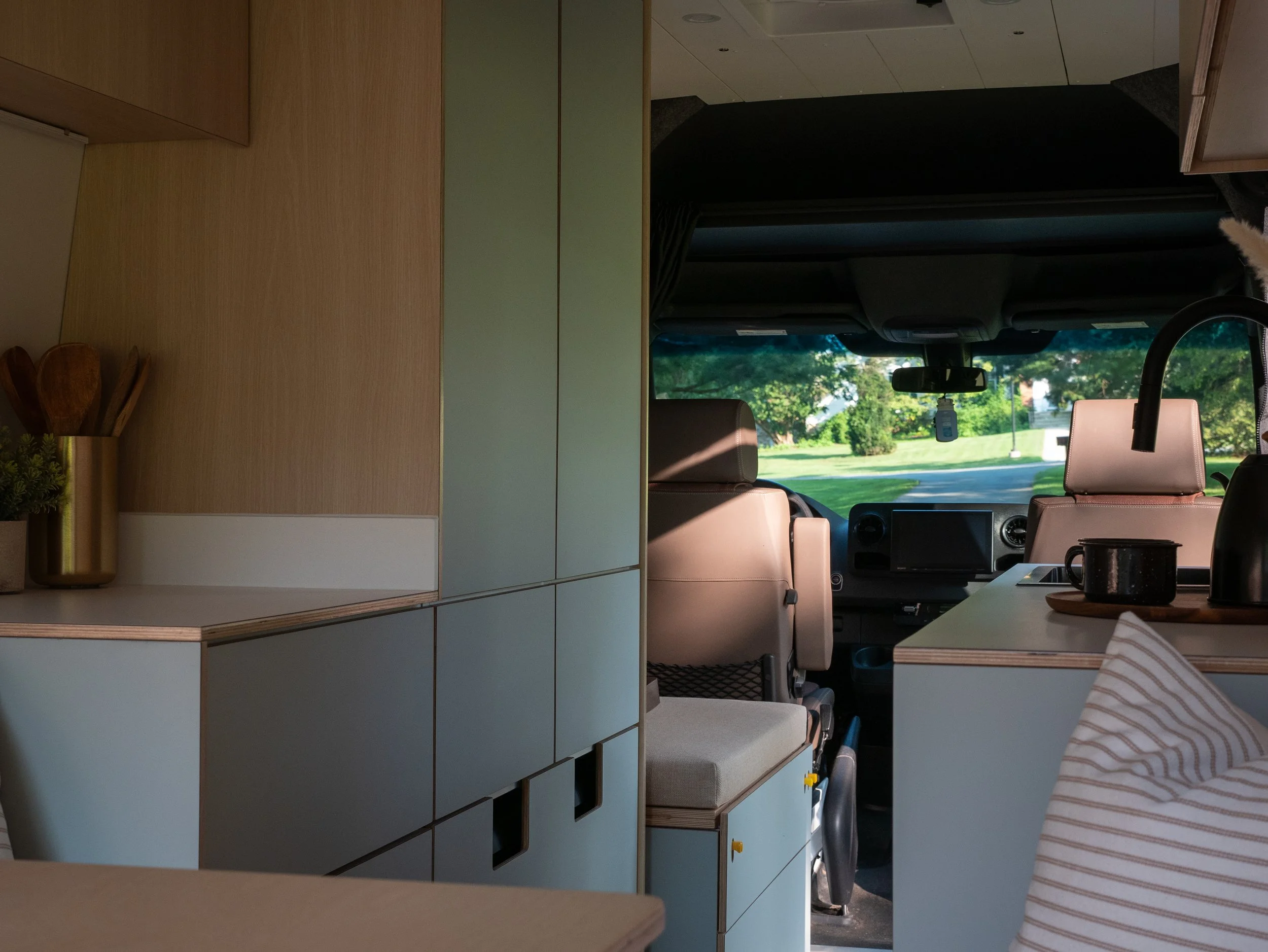 Interior of 'Aina' Mercedes Sprinter van conversion looking toward the cab — sage-green handleless lower cabinetry with routed square finger pulls, white countertop with birch plywood edge, brass utensil holder and small plant on the counter; dark br