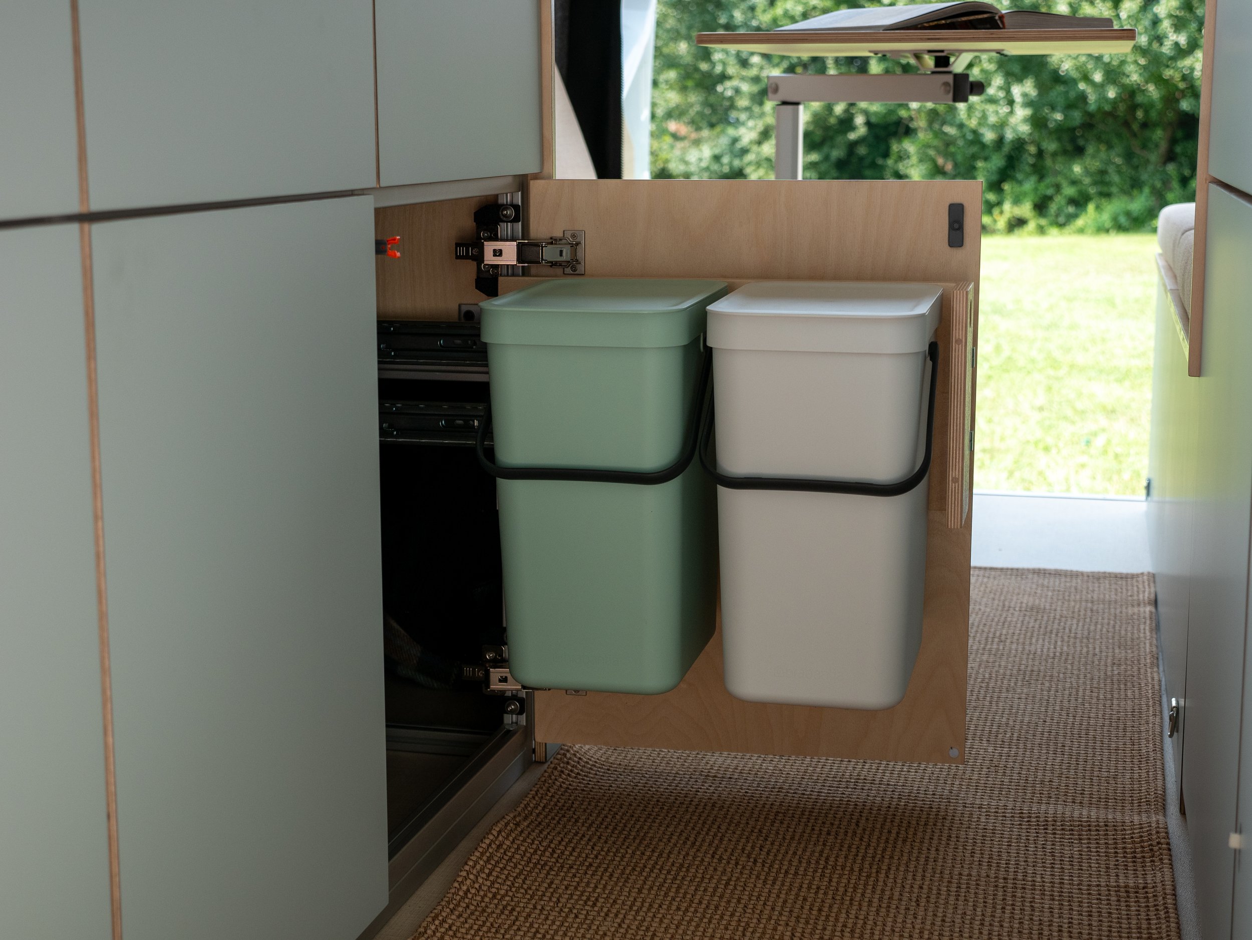 Dual waste-sorting bin pull-out in 'Aina' van conversion — sage-green and white lidded bins side by side on a birch plywood pull-out tray, heavy-duty drawer slides, cream bench cushion and Lagun folding table at right, woven jute runner on the floor,