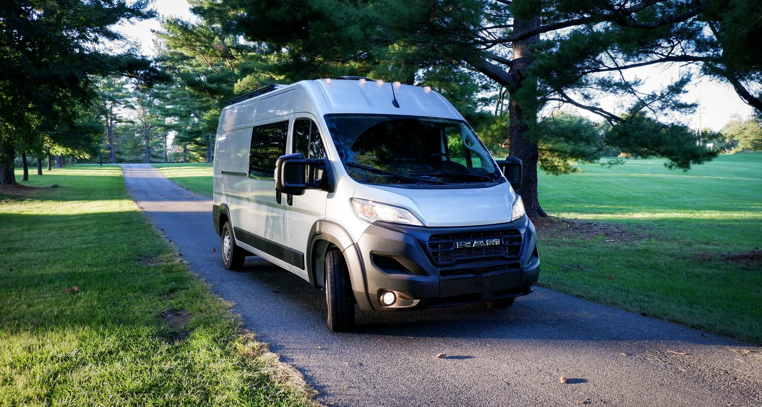 'Frejya' Ram ProMaster van conversion on a paved park path — silver body with dark grey lower trim, lush green trees and manicured lawn; a custom campervan build featuring a loft sleeping platform above a rear gear garage.