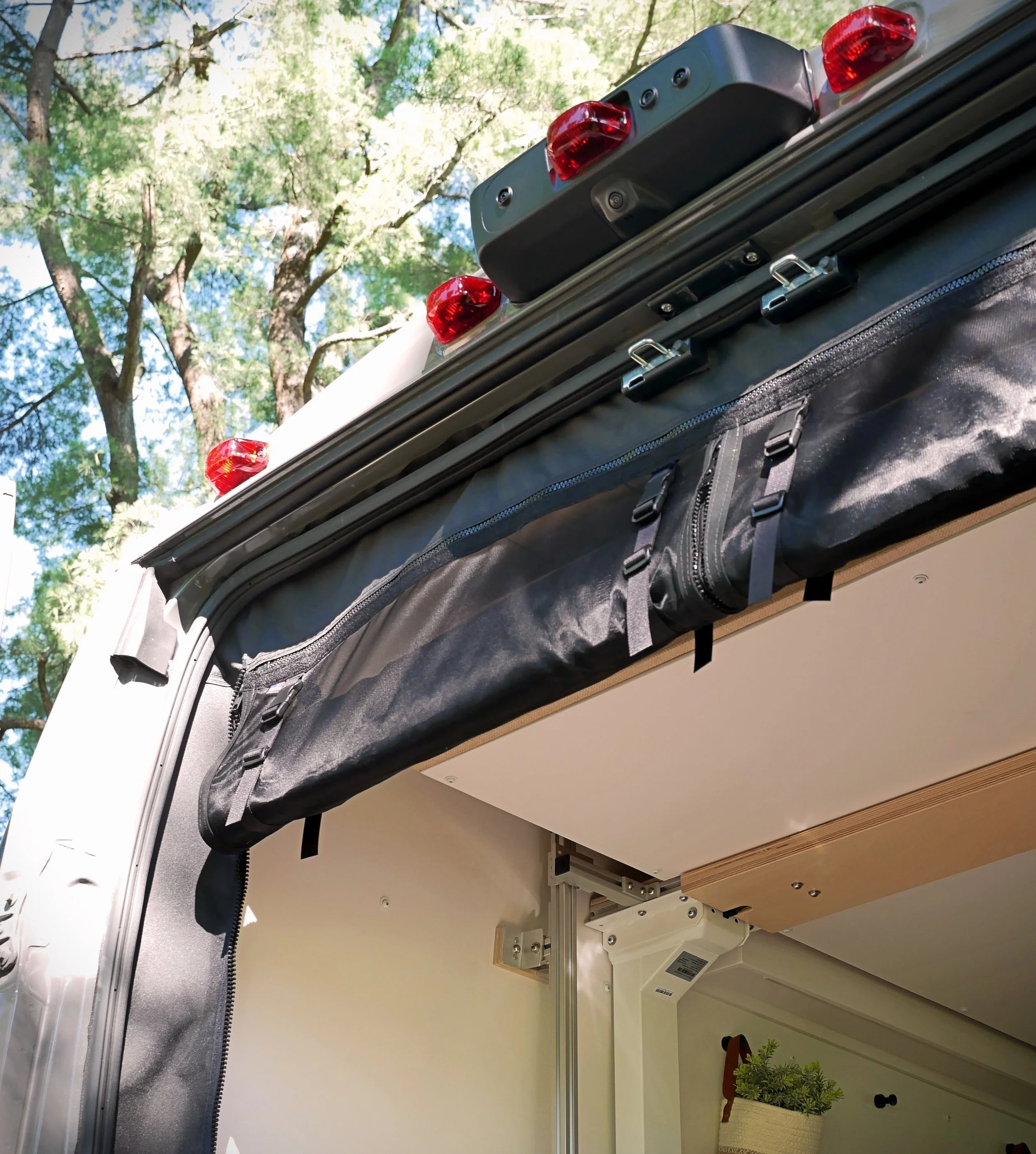 Bug screen detail on 'Bjorn' Ram ProMaster van conversion — black bug screens with red clearance lights and backup camera on the bracket, rolled black fabric secured with straps, photographed from below against a green pine tree canopy.