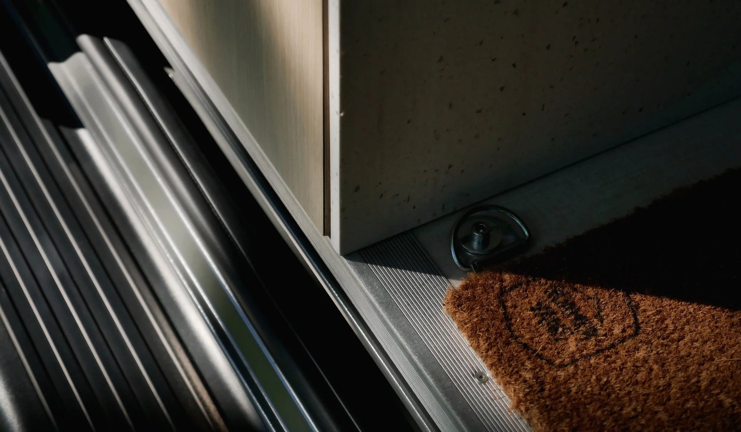 Entry threshold in 'Bjorn' Ram ProMaster van conversion — aluminum sliding door track and ribbed door sill, chrome D-ring anchor hardware at the base of the white quartz kitchen panel, branded coir entry mat in dramatic shadow.