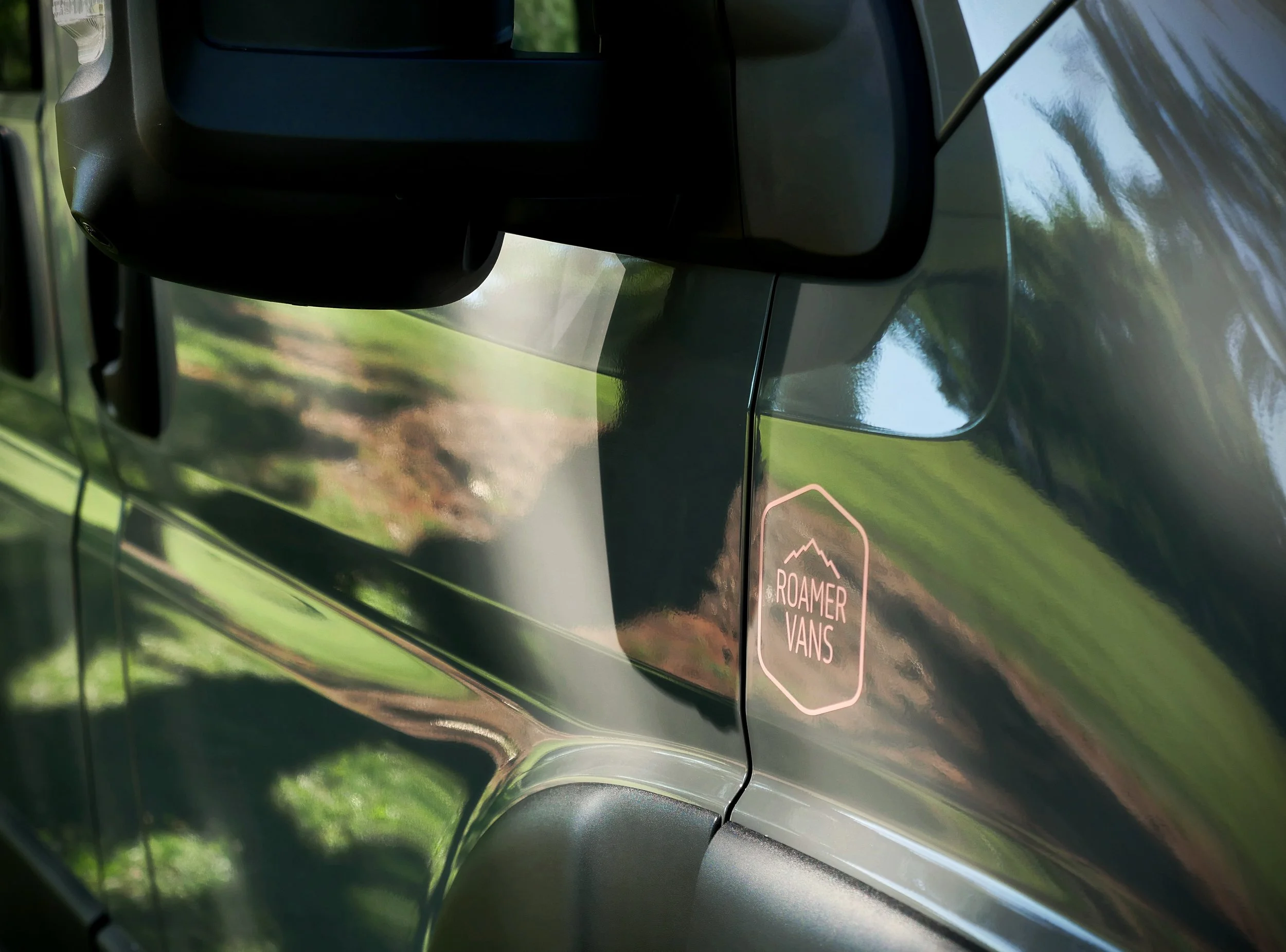Roamer Vans branding detail on 'Bjorn' van conversion — salmon pink octagonal logo badge affixed to the grey Ram ProMaster body panel near the side mirror, lush green landscape reflected across the van's polished finish.