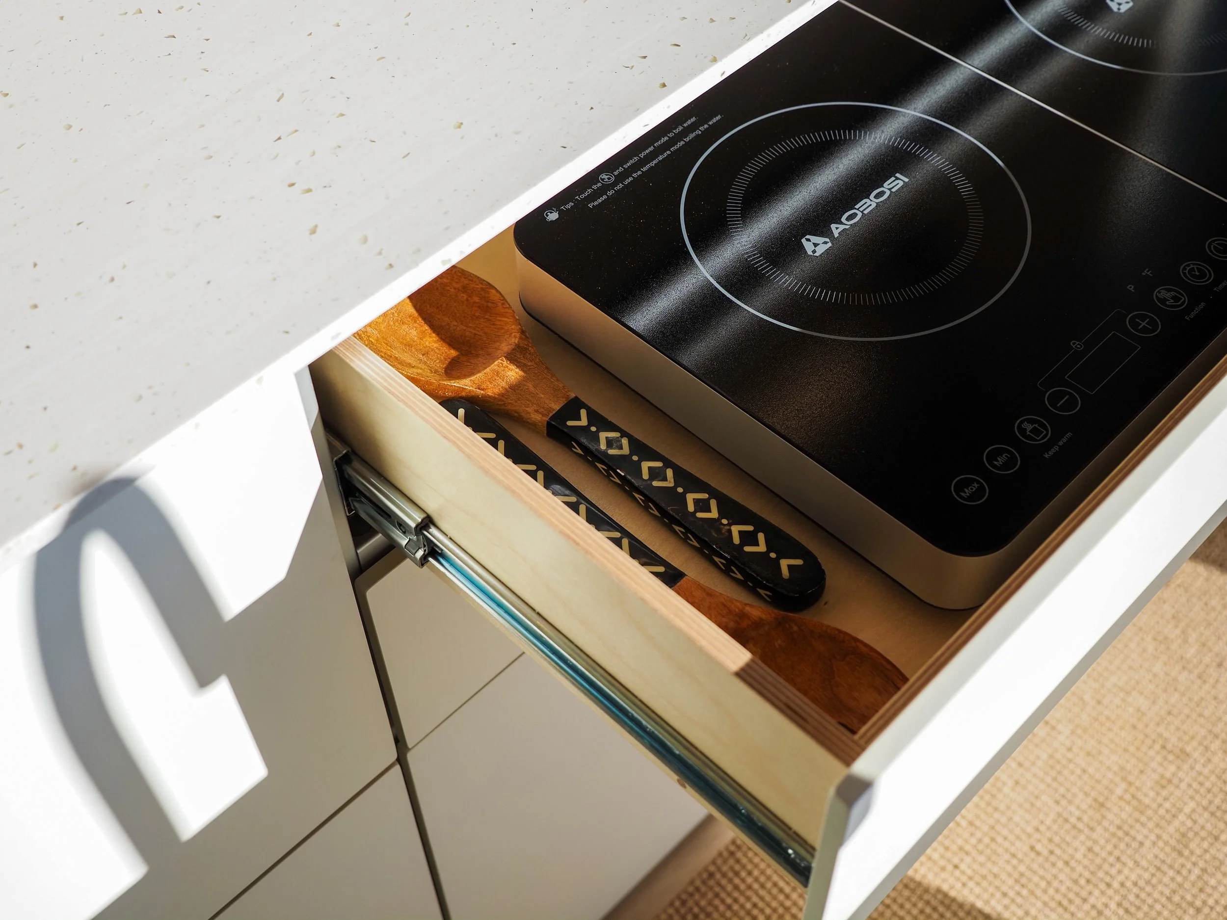Hidden pull-out induction cooktop in 'Frida' Ram ProMaster van conversion — black dual-burner unit stored in a birch plywood drawer on full-extension slides beneath the white solid surface countertop, wooden cooking utensils alongside, woven floor ma