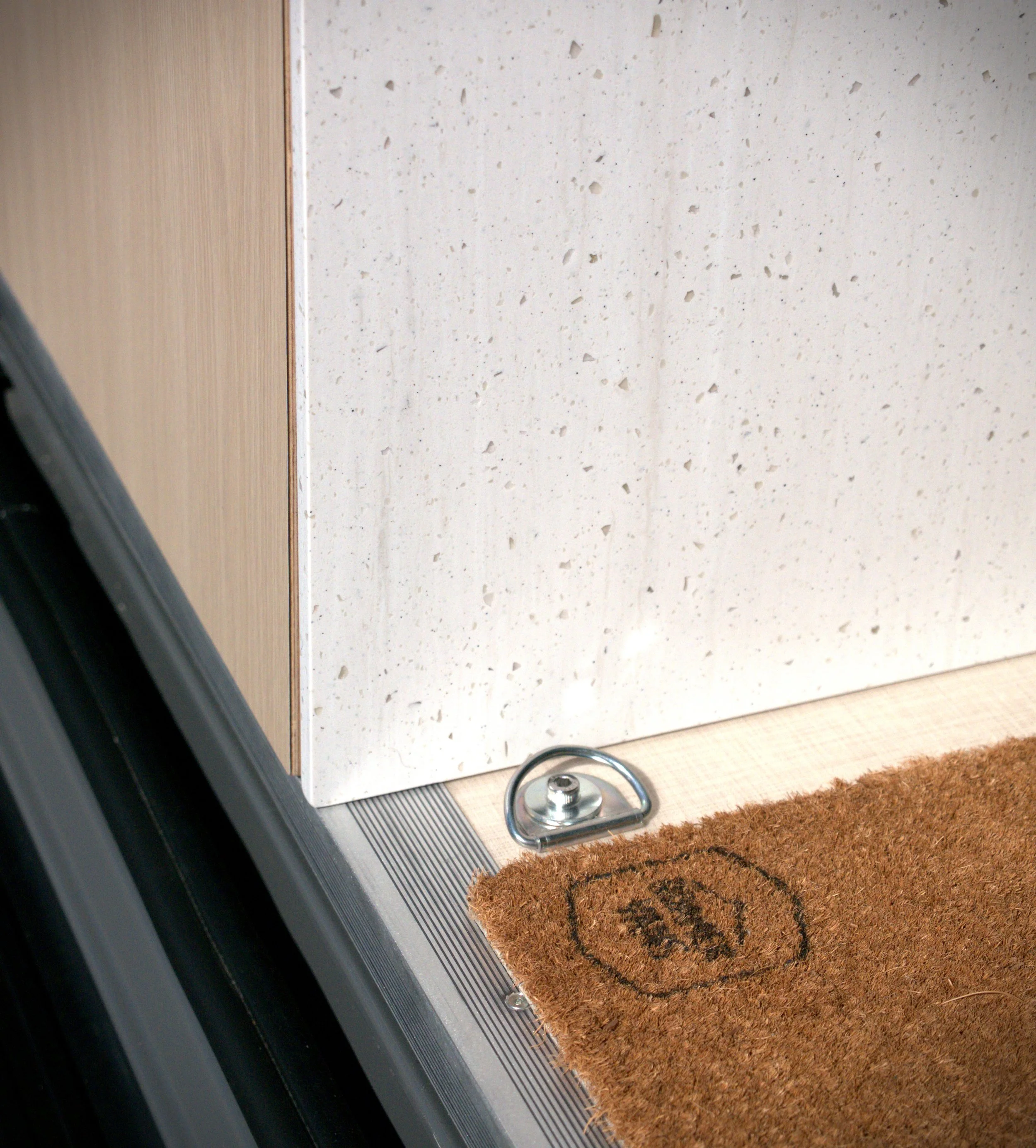 Van conversion entry threshold detail in 'Bjorn' — stainless steel D-ring tie-down anchor flush-mounted in the birch plywood floor beside the sliding door, branded Roamer Vans coir welcome mat, and white waterfall countertop above.