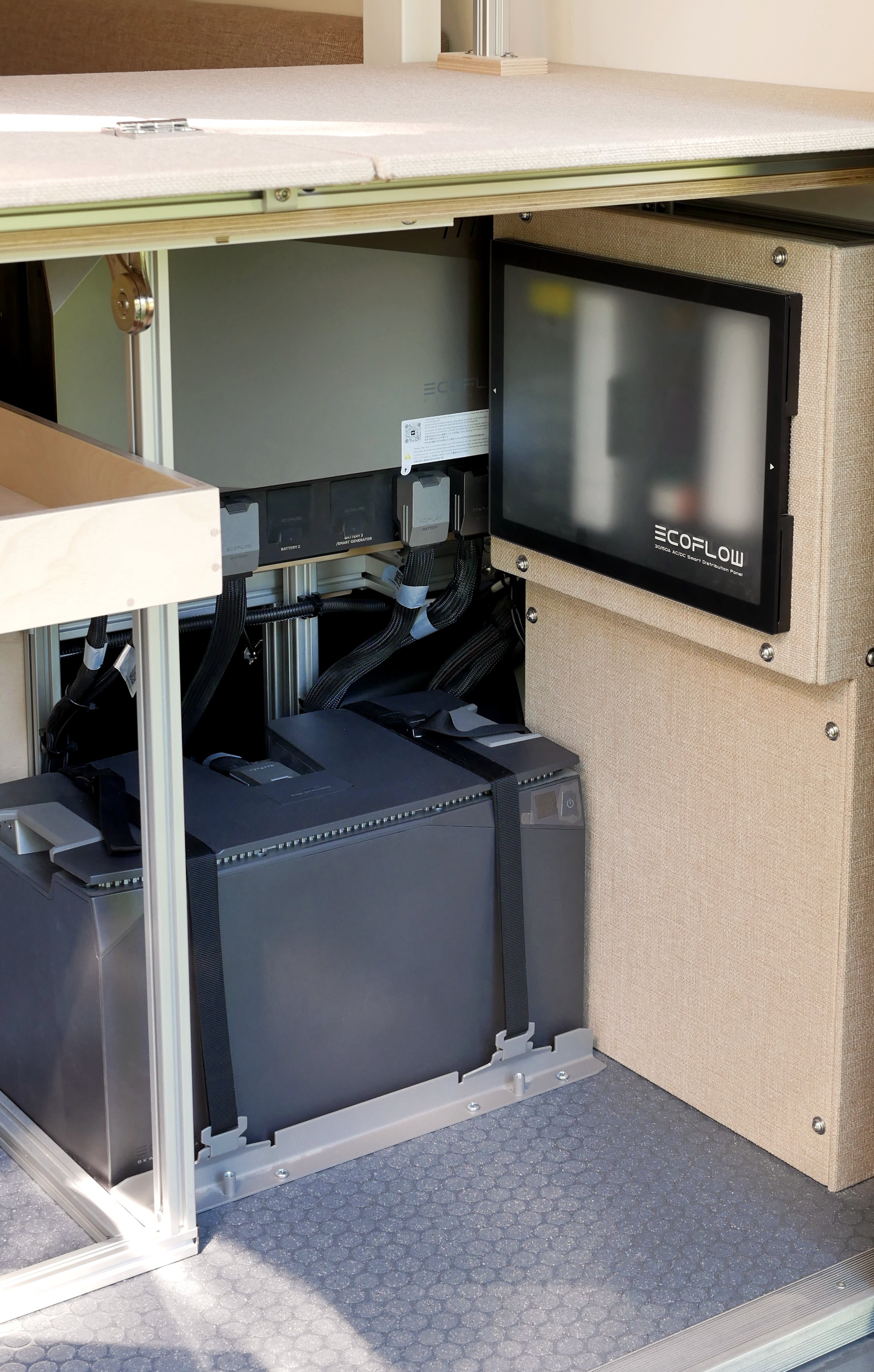 Van conversion electrical system in 'Bjorn' — EcoFlow lithium battery in an aluminum-framed rear compartment with rubber diamond-plate floor mat, EcoFlow distribution panel on the beige wall panel, wiring and power components visible above.