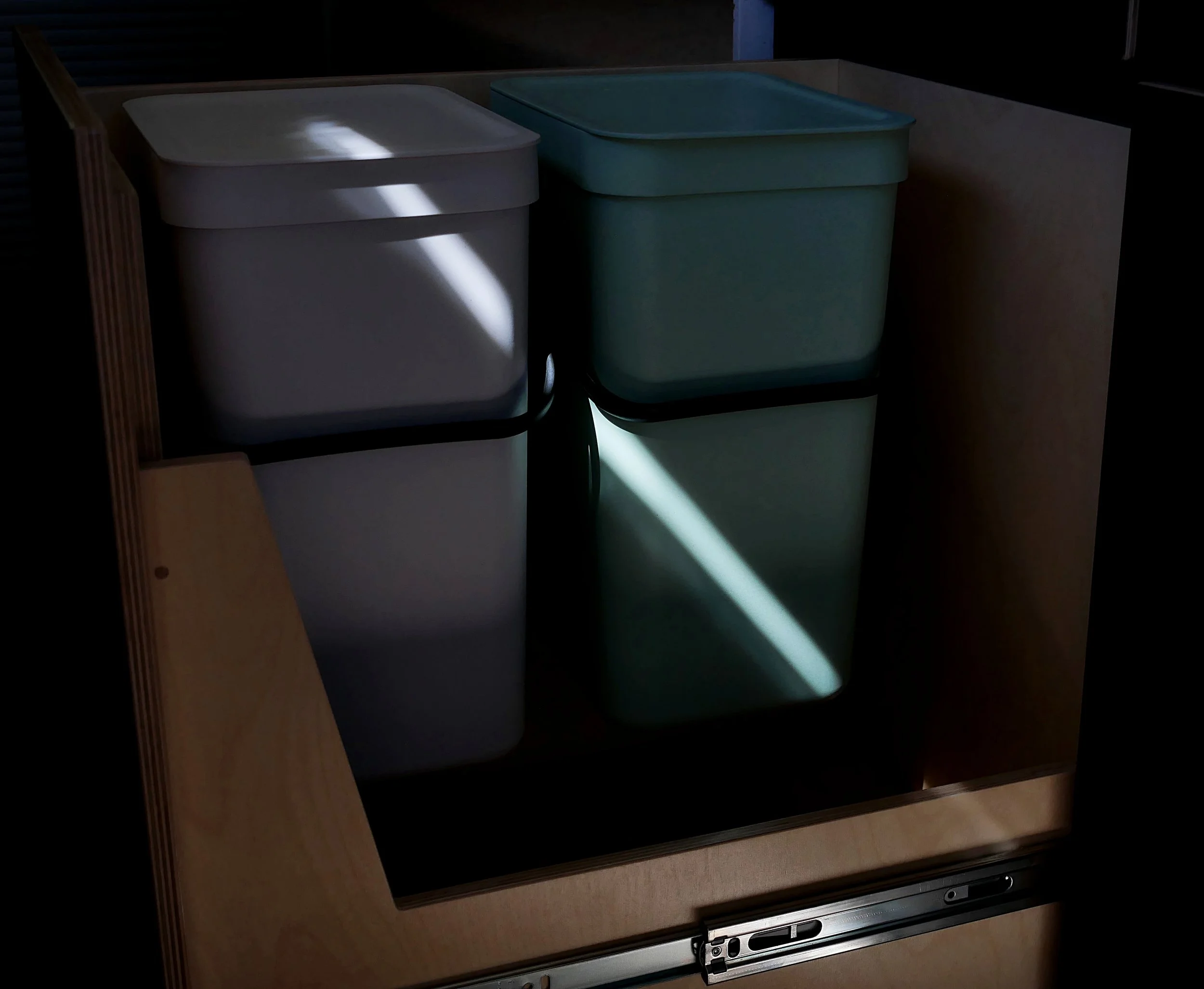 Pull-out waste bin system in 'Bjorn' van conversion — birch plywood drawer on full-extension slides holding a white and a teal bin for trash and recycling, dramatic shadow-and-light composition.
