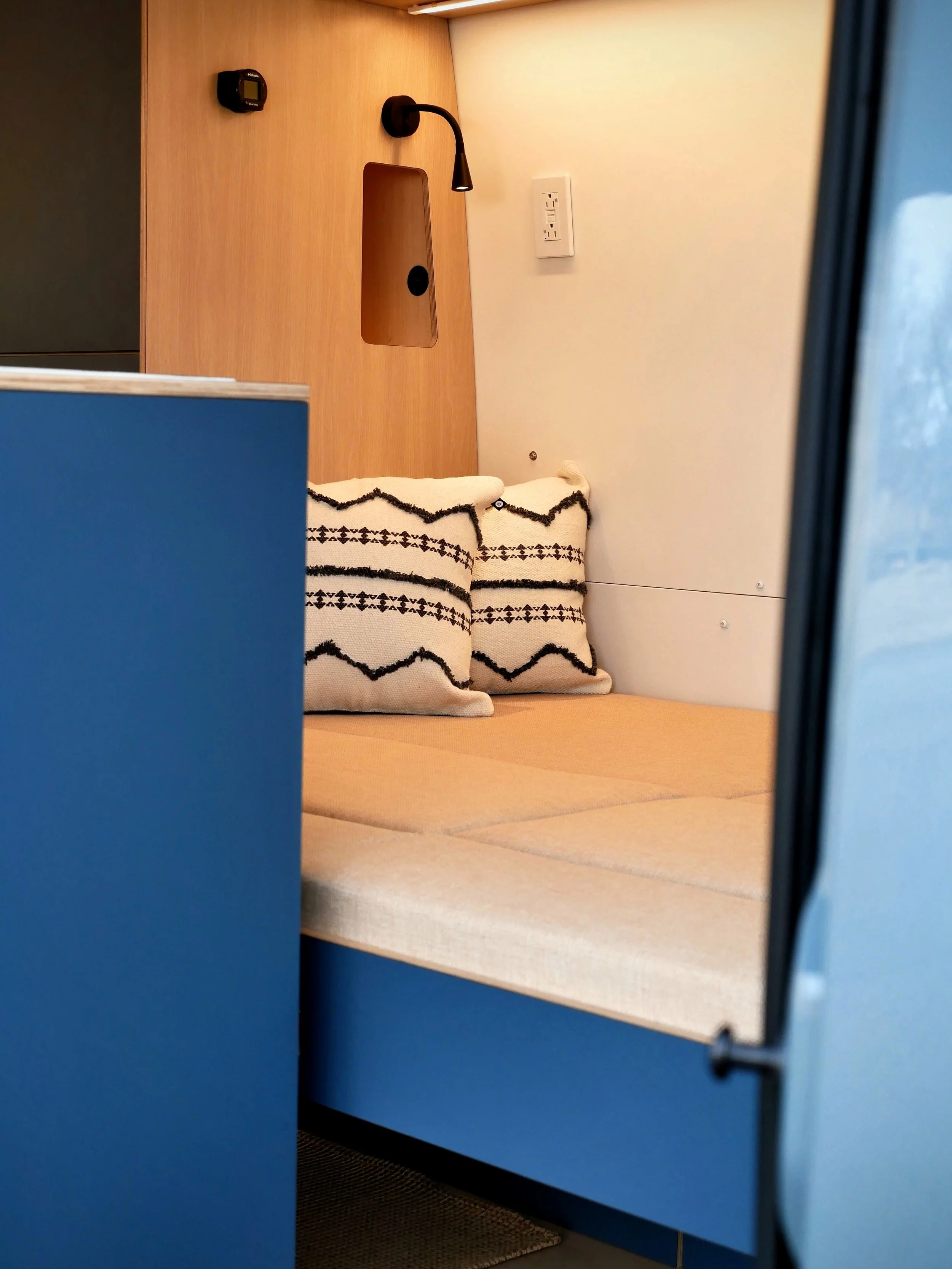 Sleeping area of 'Nordlys' conversion viewed through the open sliding door — beige seat and bed cushions with geometric cushions, black reading lamp, recessed charging nook, LED strip lighting