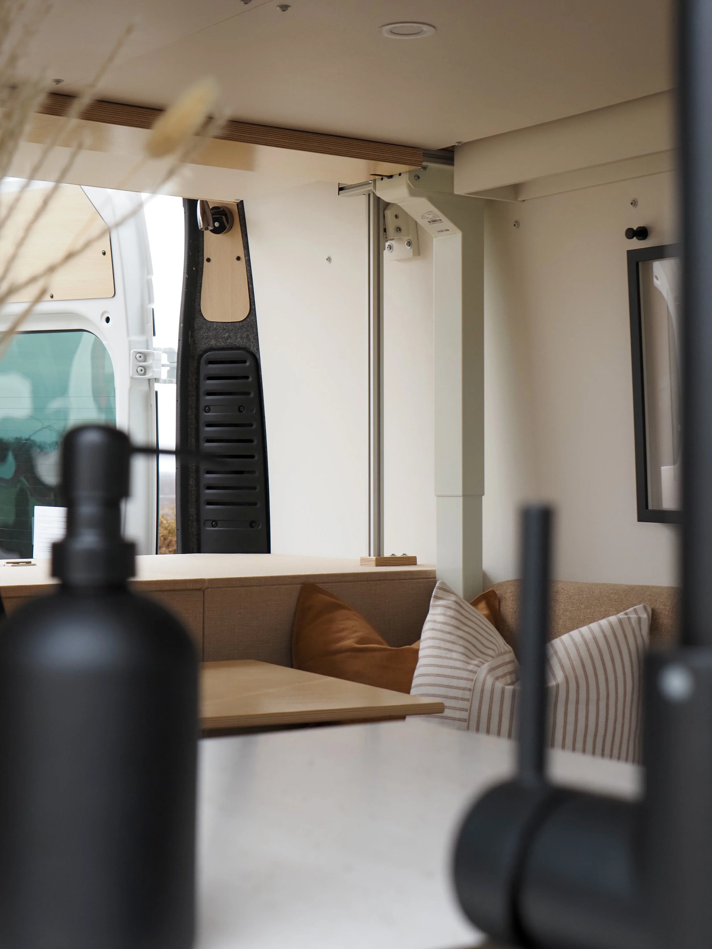 Looking through the kitchen of 'Frida' Ram ProMaster van conversion — black sink basin and gooseneck faucet in soft-focus foreground, dried pampas stems at the upper edge; U-shaped lounge with terracotta pillow and striped cushions beyond, chrome sho