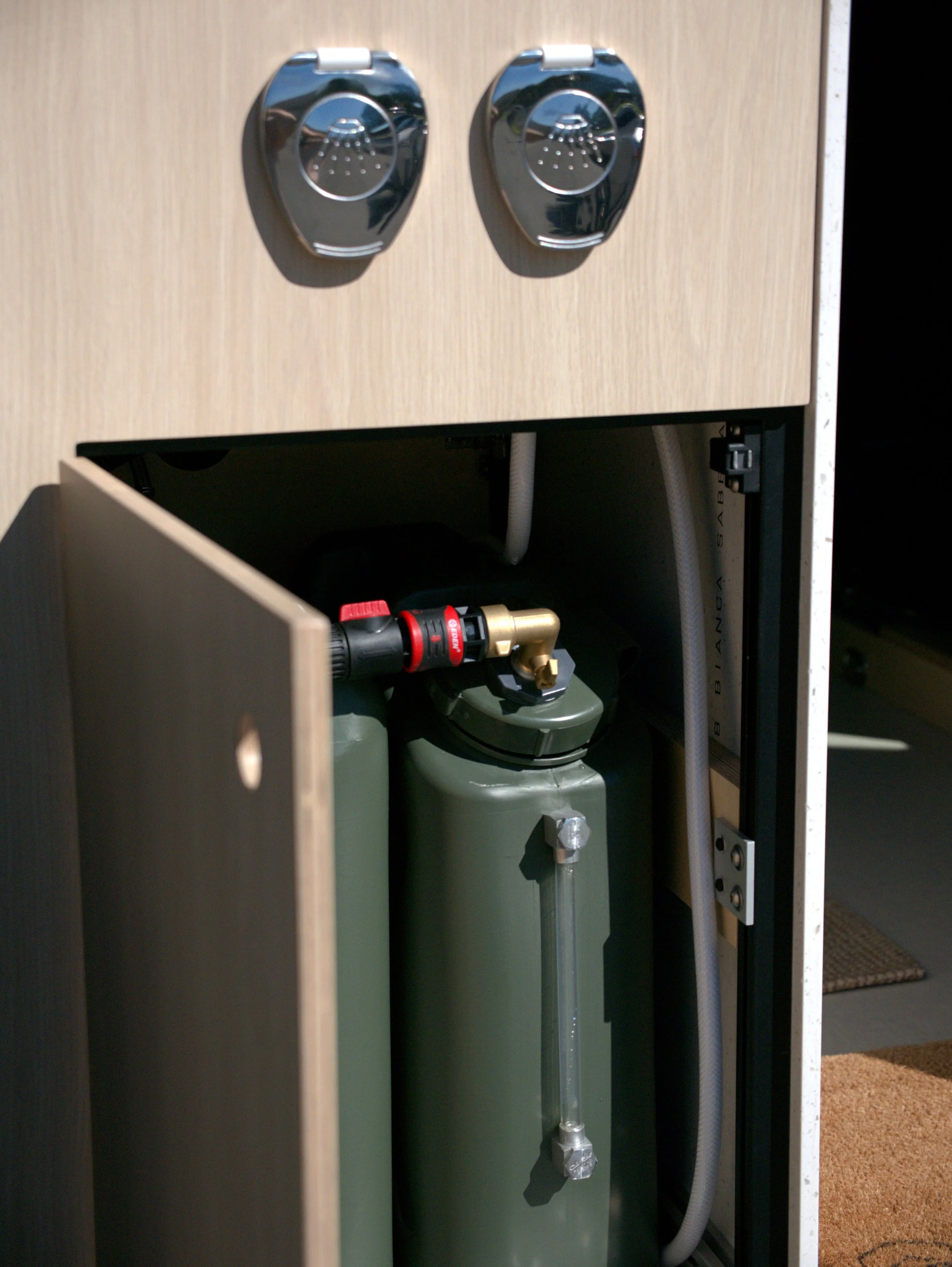 Fresh water system in 'Bjorn' Ram ProMaster van conversion — lower door of the entry kitchen module open to reveal twin olive-green water tanks with brass and quick-connect plumbing fittings; birch cabinet face with chrome Southco latches visible abo