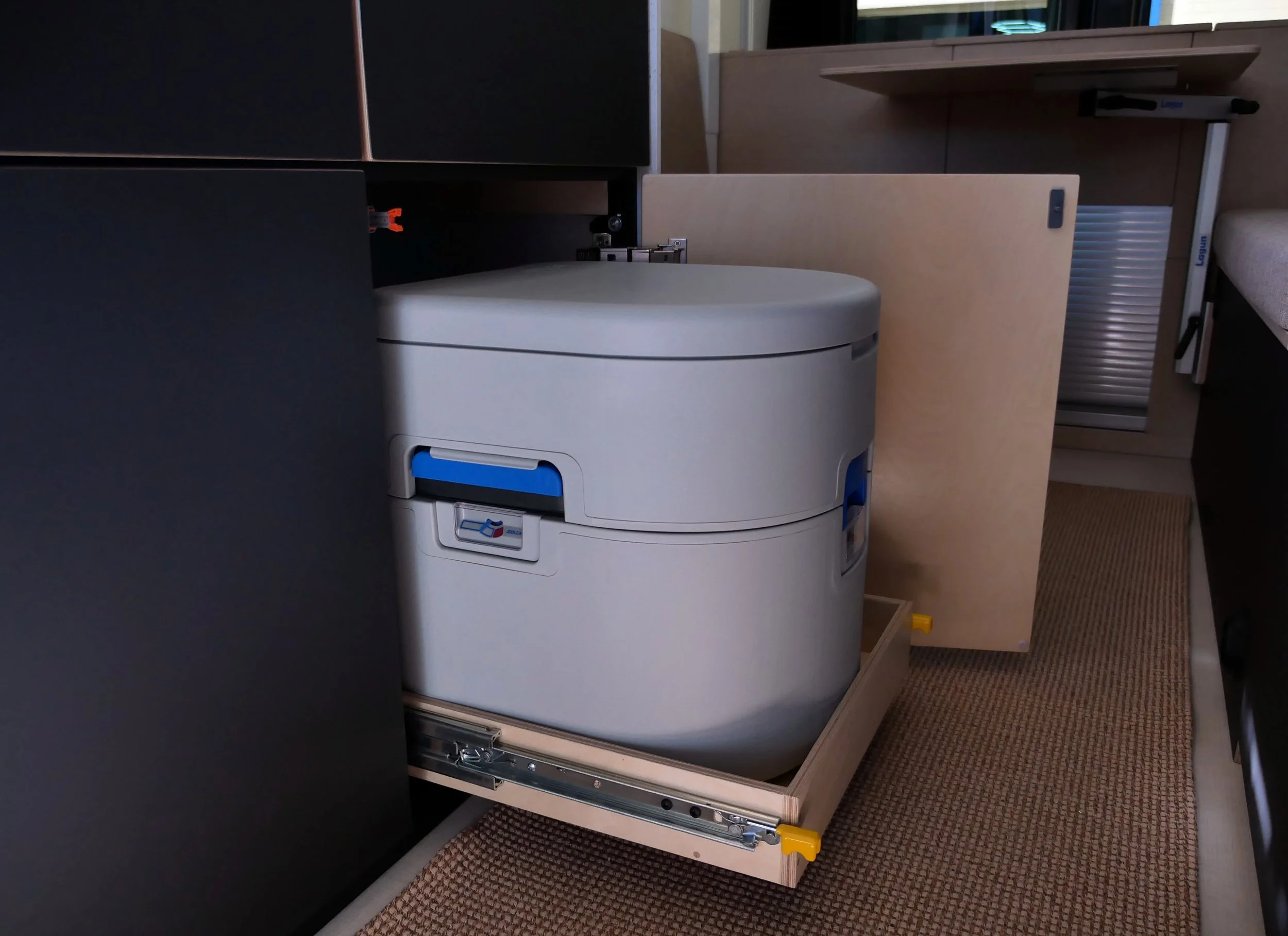 Hidden separator toilet in 'Bjorn' van conversion — Joolca GottaGo on a birch plywood slide tray pulled from concealed under-bench storage, yellow safety latch visible, bench cushion and Lagun table arm in the background.