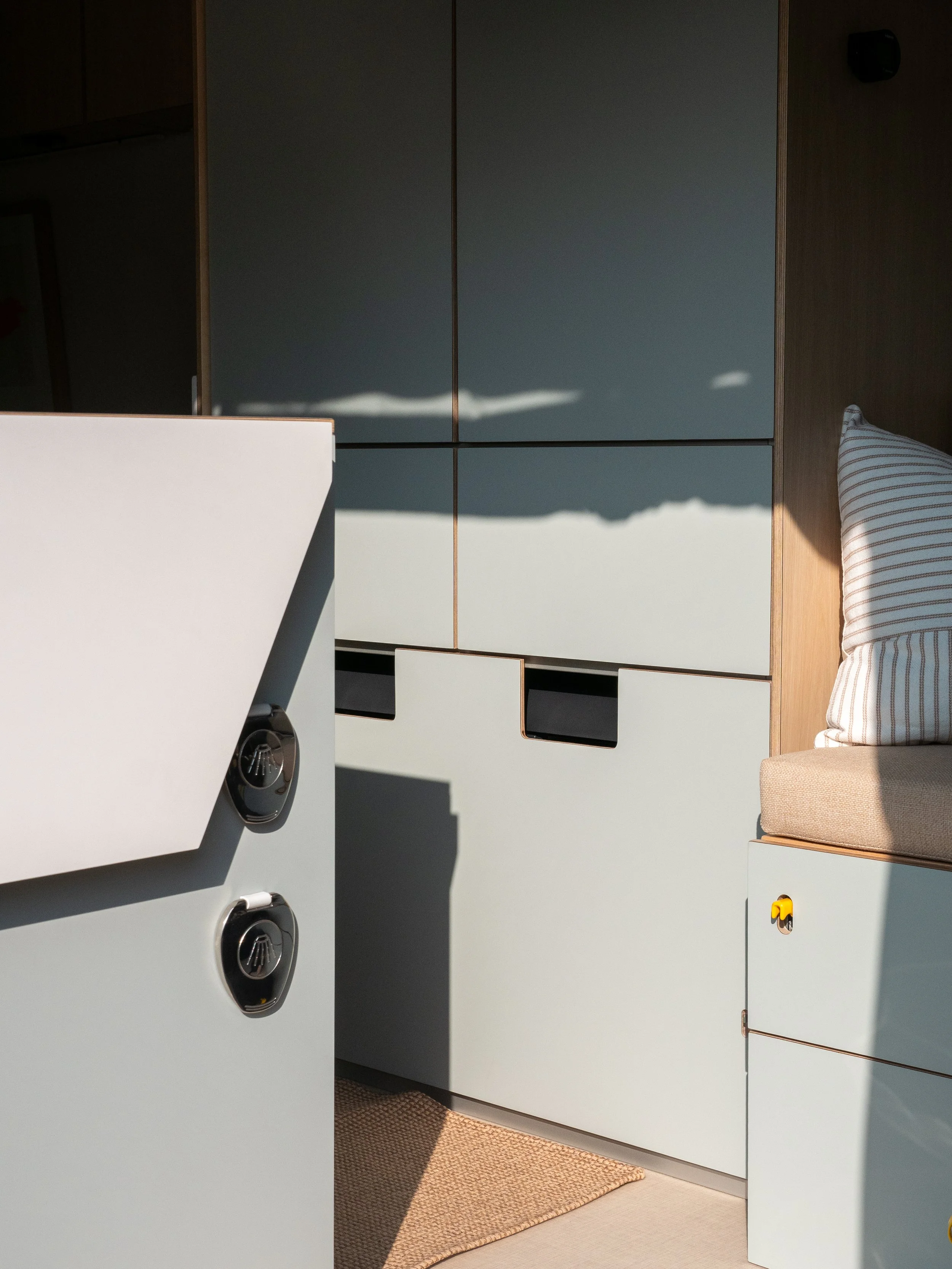 Galley cabinetry in 'Aina' van conversion — sage-green flat-front panels with finger-pull cutouts for drawer and a hinged countertop extension and shower outlets; bench seat with striped pillow and gold accent detail in warm raking sunlight.