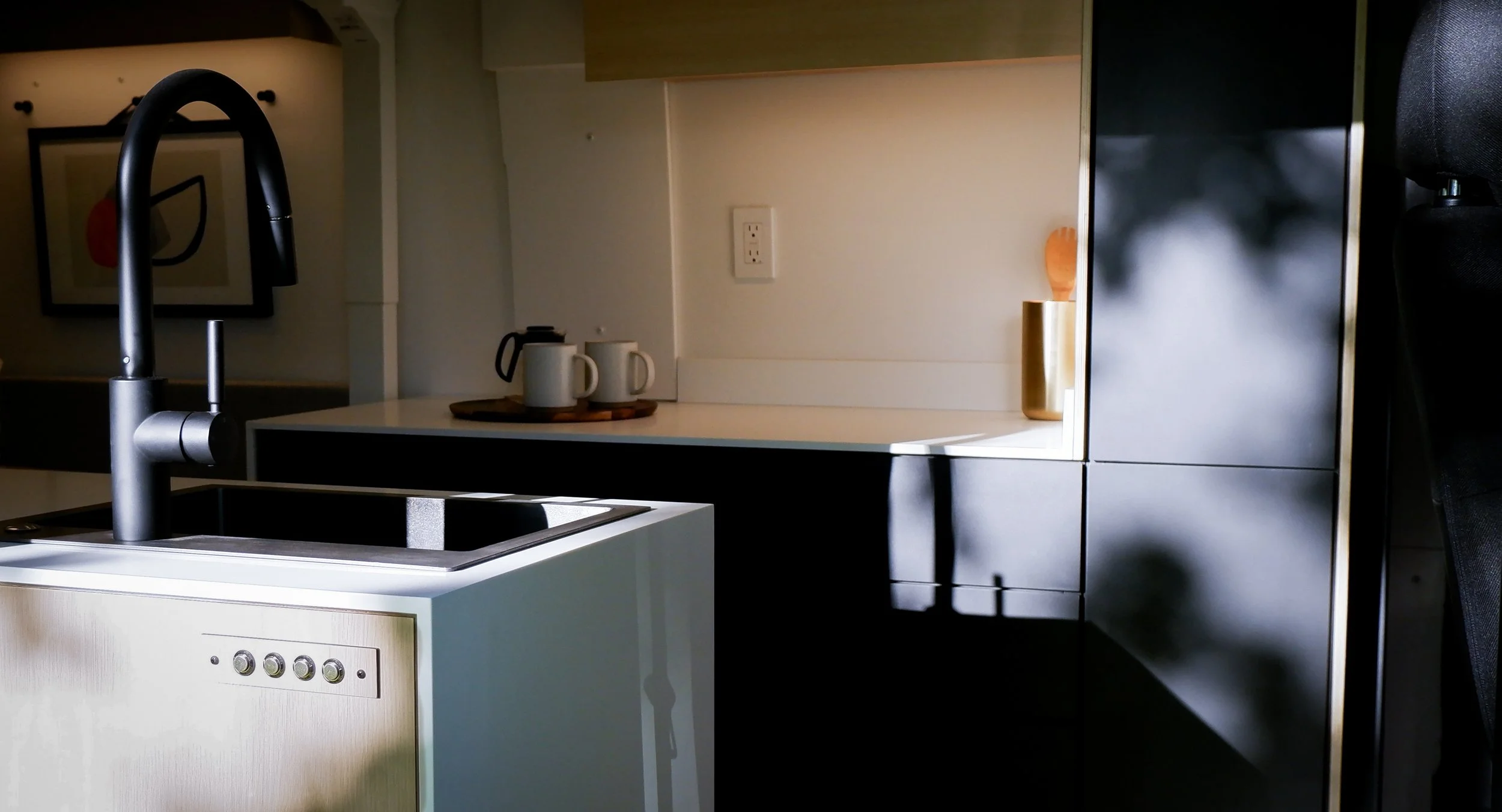 Custom van conversion kitchen in 'Frejya' — distinctive dark interior with black handleless cabinet faces, black gooseneck faucet, white waterfall countertop, pour-over kettle and mugs on a wooden tray, small brass utensil holder on the counter