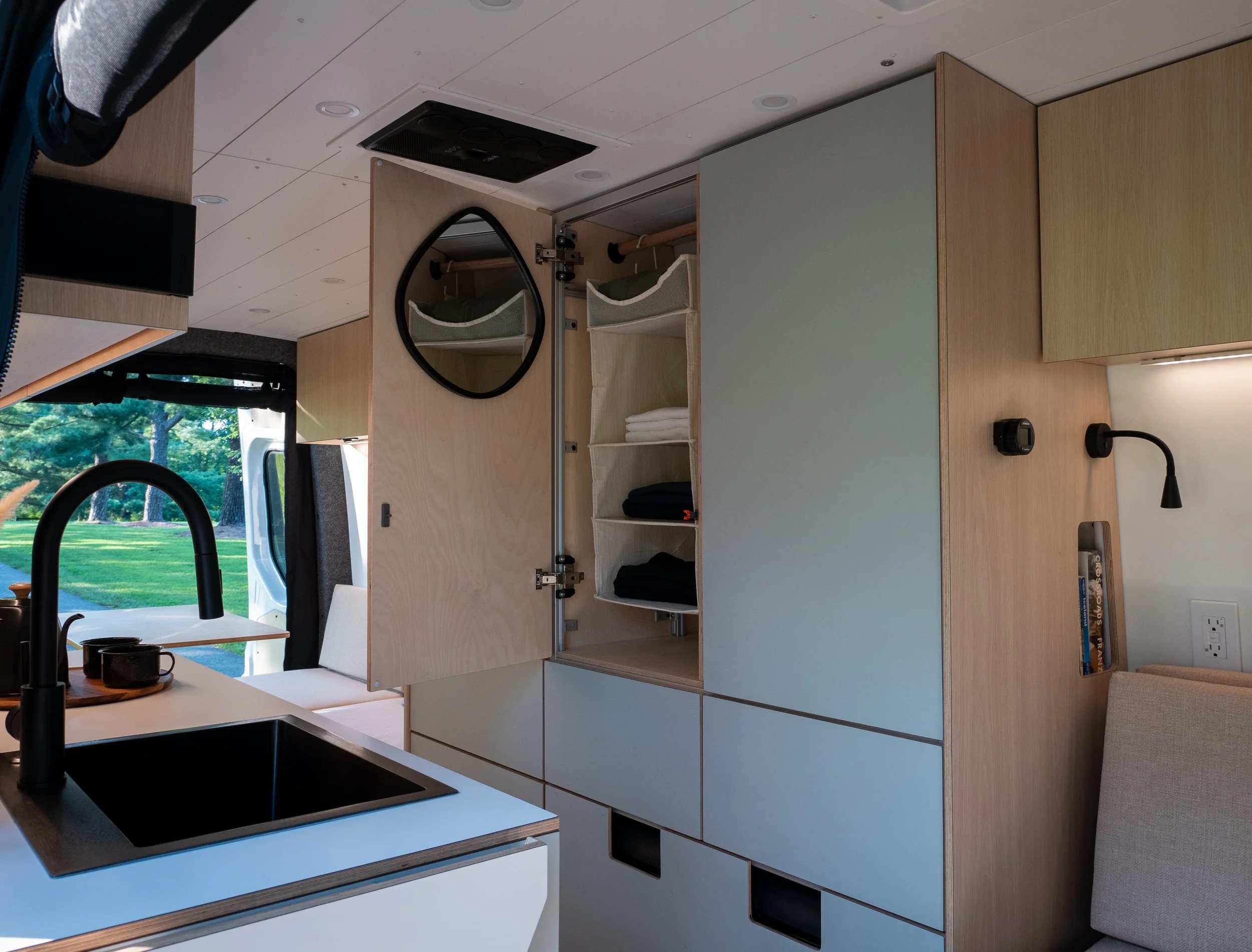 Interior of 'Aina' Sprinter van conversion looking toward the cab from the sleeping area — tall sage-green wardrobe open with canvas pocket organizer, folded towels, and clothing inside; round black-framed mirror mounted above; van conversion kitchen