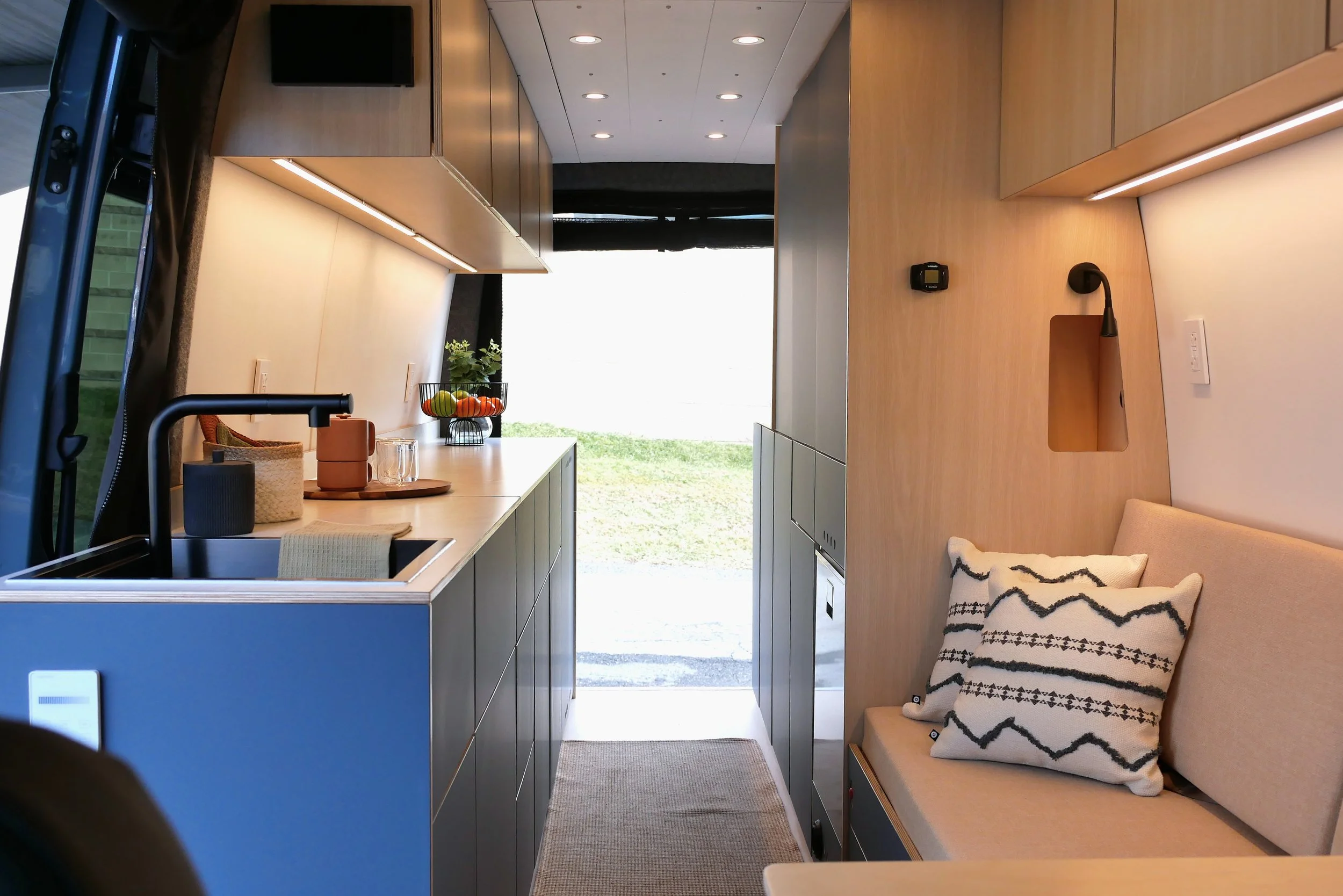 Interior of 'Nordlys' looking toward the open sliding door — galley kitchen with blue cabinets and black sink on left, bench seat with geometric cushion and wall-mounted reading light on right