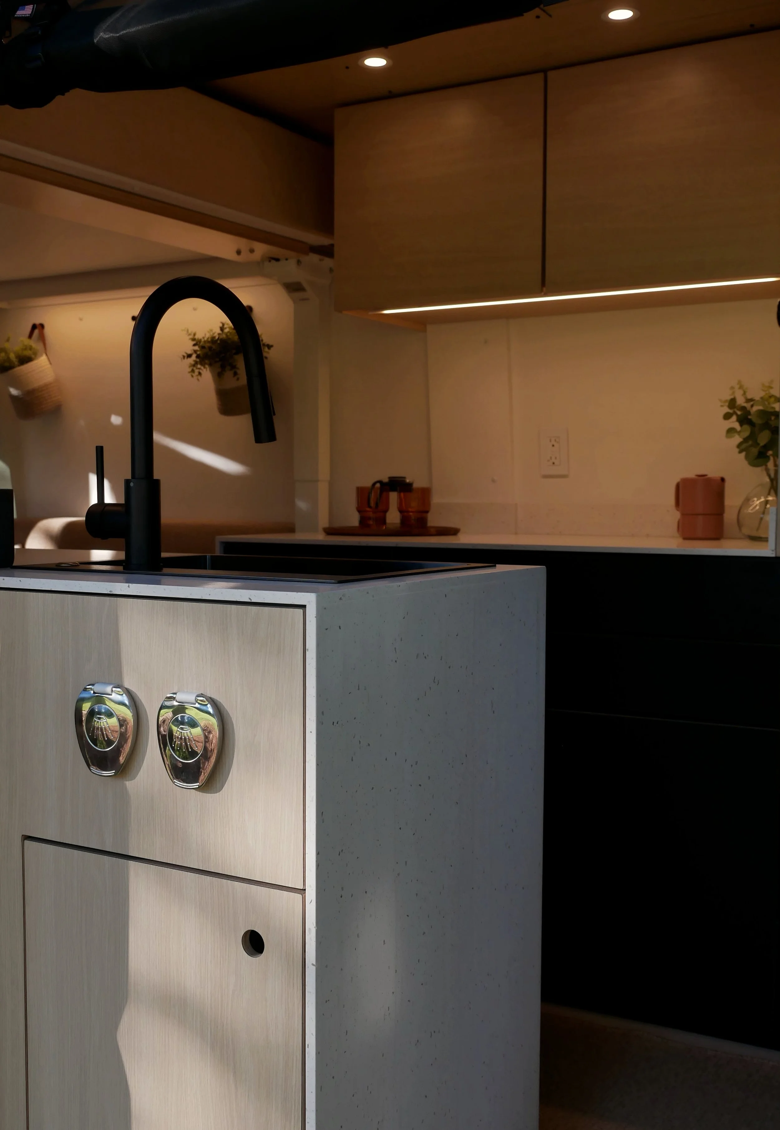 Evening interior of 'Bjorn' Ram ProMaster van conversion — entry kitchen module with birch cabinet face and chrome latches in the foreground, galley kitchen beyond with white countertop, warm under-cabinet LED strip, terracotta accent vessels, and fl
